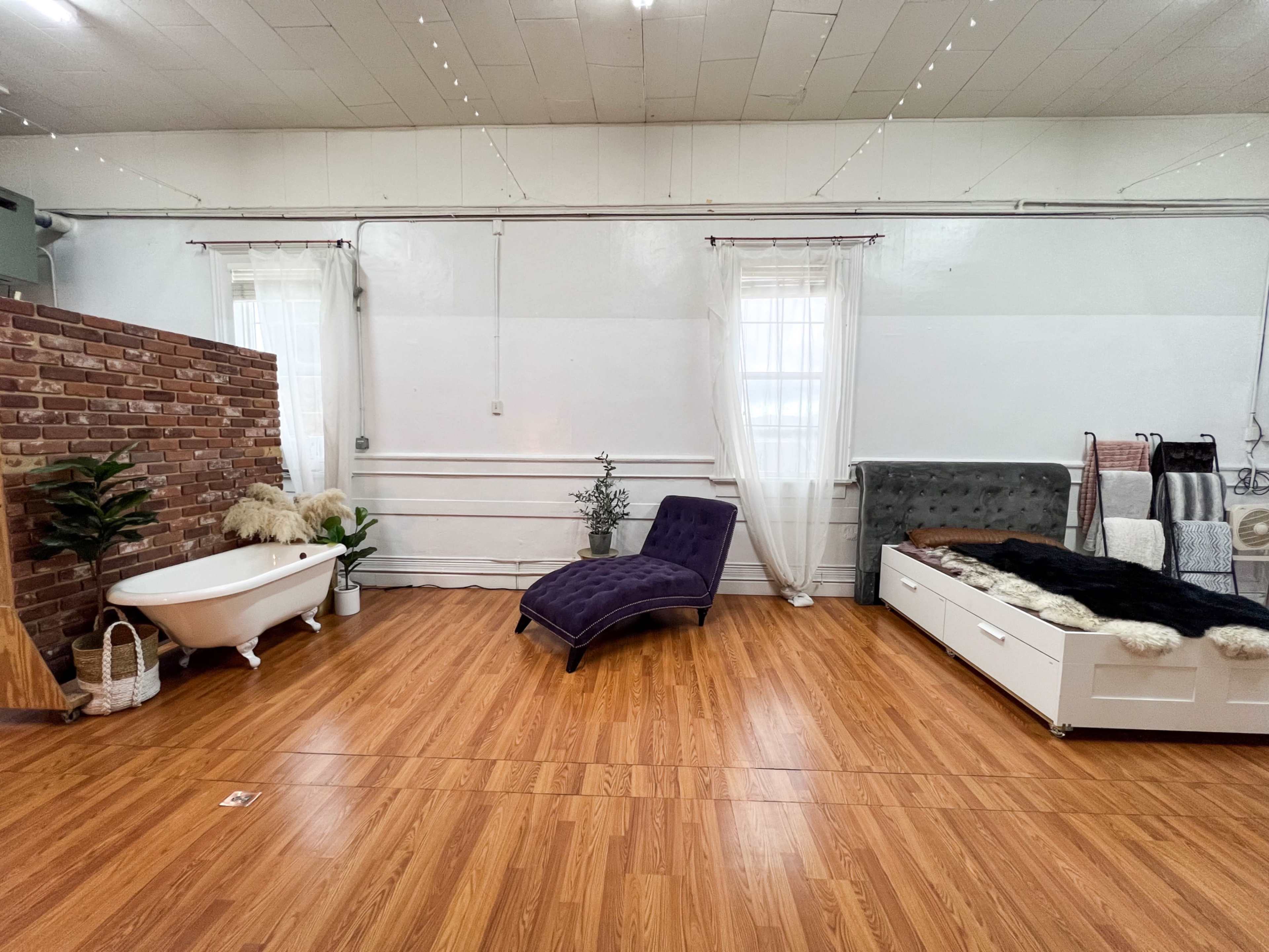 The image shows a spacious room with wooden flooring, featuring a vintage bathtub, a purple chaise lounge, and a bed with a tufted headboard.