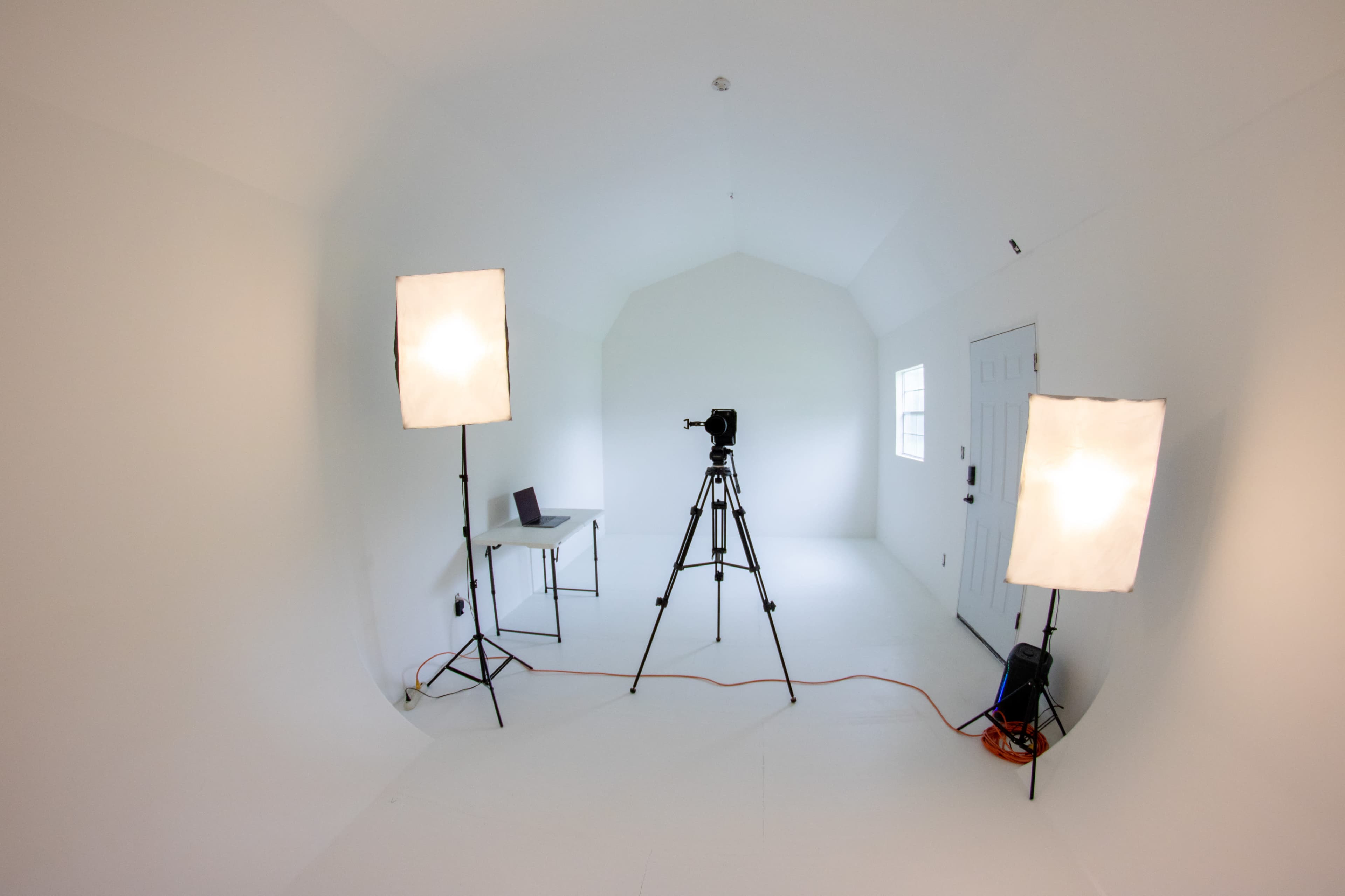 A minimalist studio space features a camera on a tripod, two lighting setups, a small table, and a door to the outside.