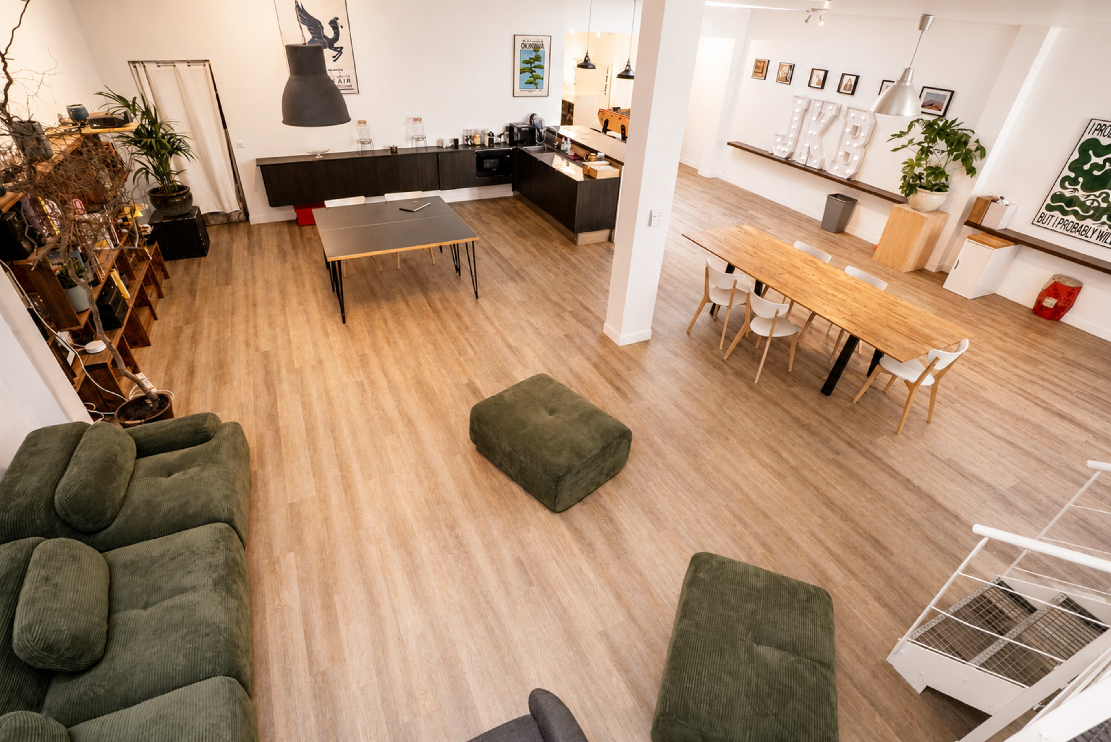 The image shows a spacious, minimalistic interior with a large table, seating areas, and a kitchen counter, all arranged in a modern workspace.