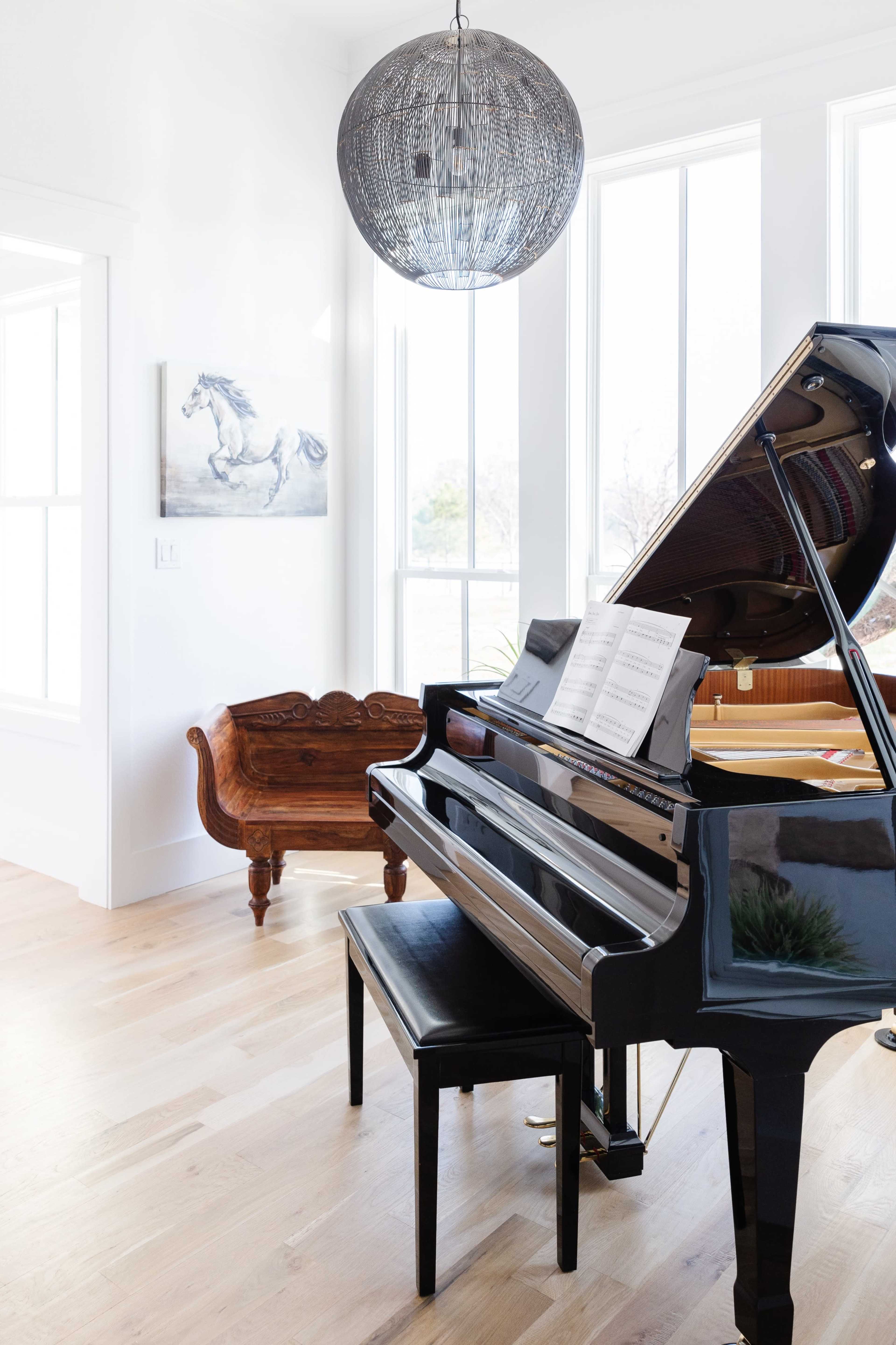A black piano with sheet music sits beside a wooden bench in a bright room featuring large windows and a decorative light fixture.