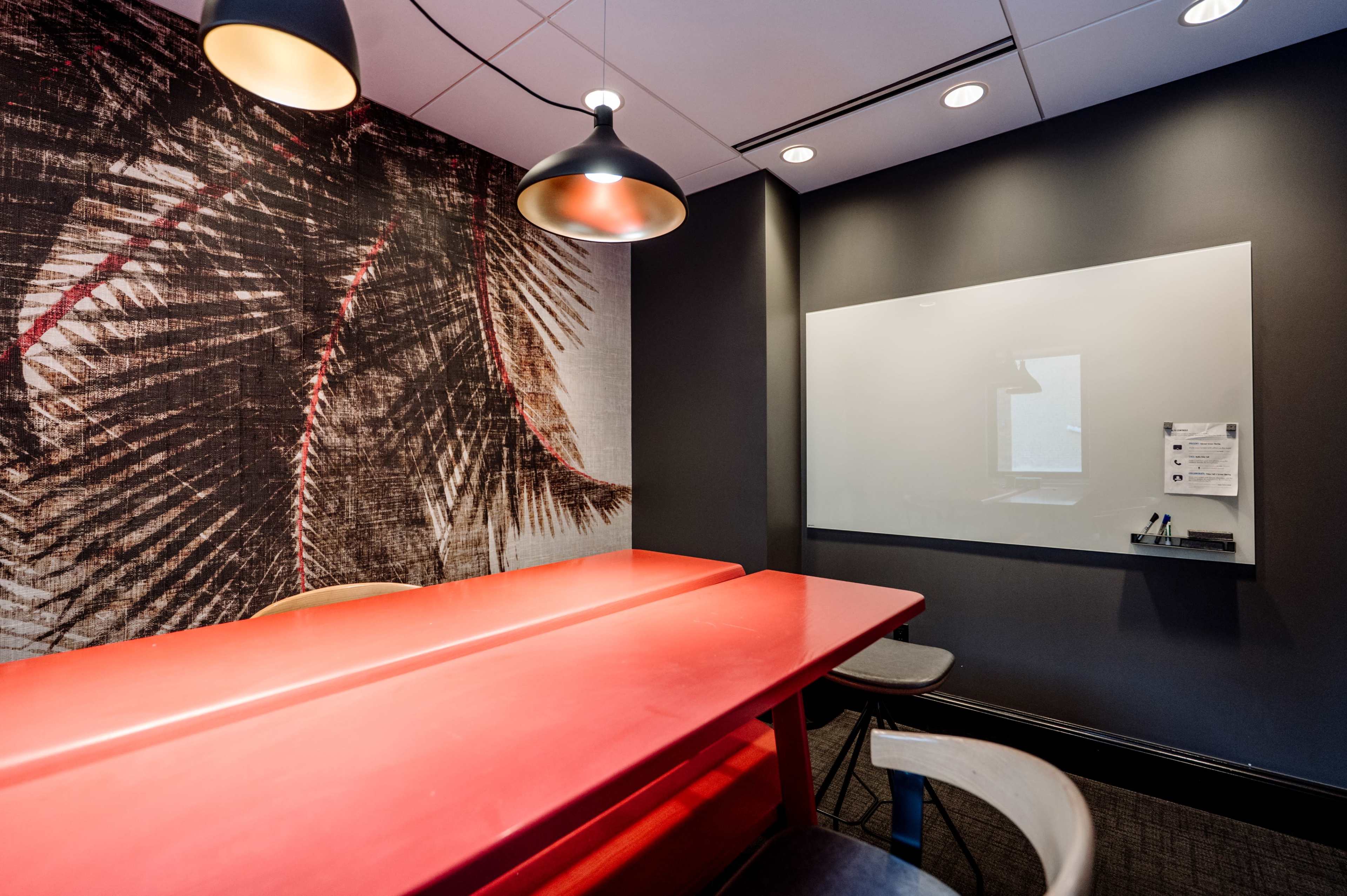A meeting room features a red table, minimalist chairs, and a whiteboard on the wall, with a palm leaf design wallpaper.