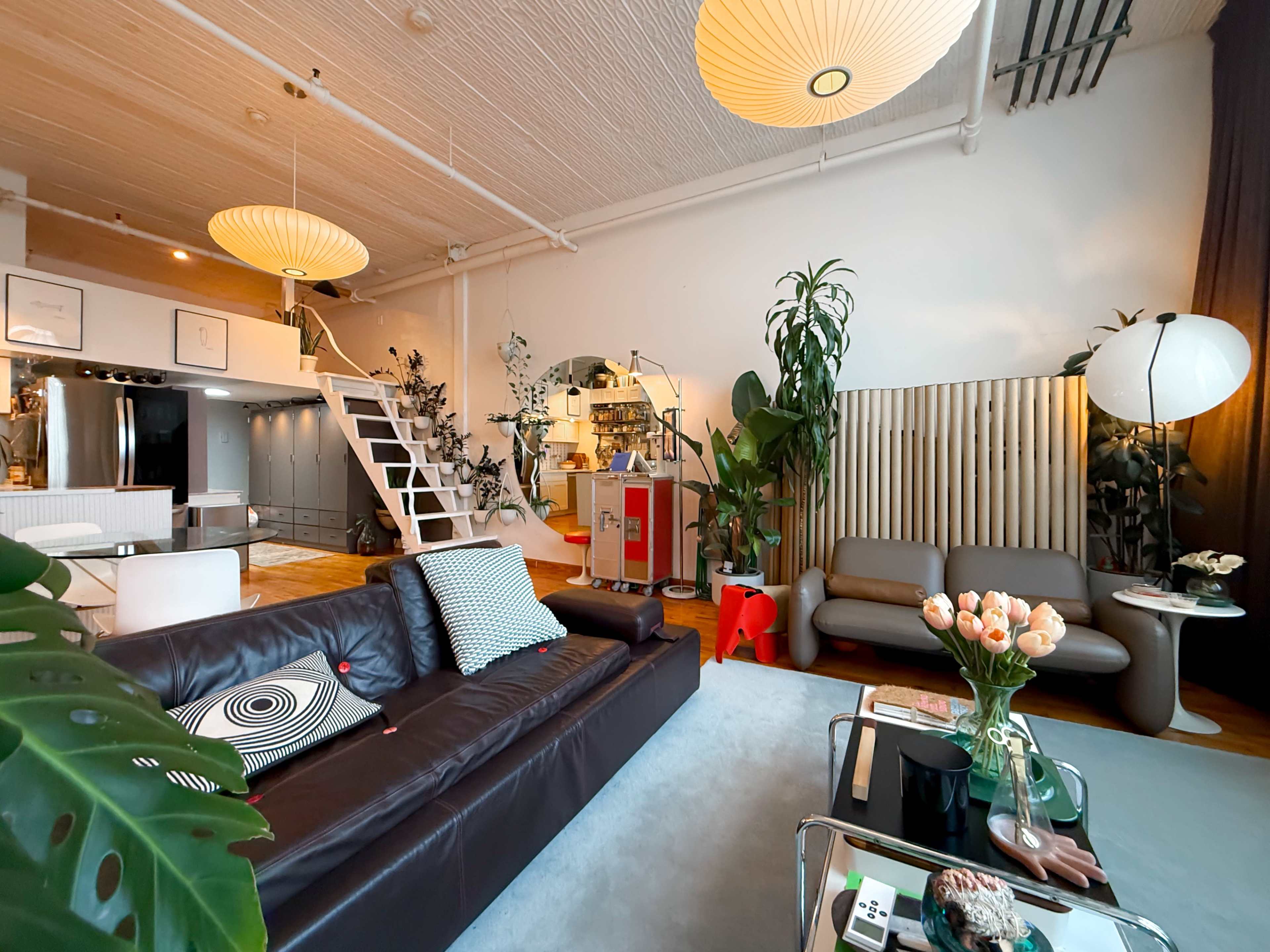 The image shows a modern living room featuring a dark sofa, decorative plants, pendant lighting, and a staircase in the background.