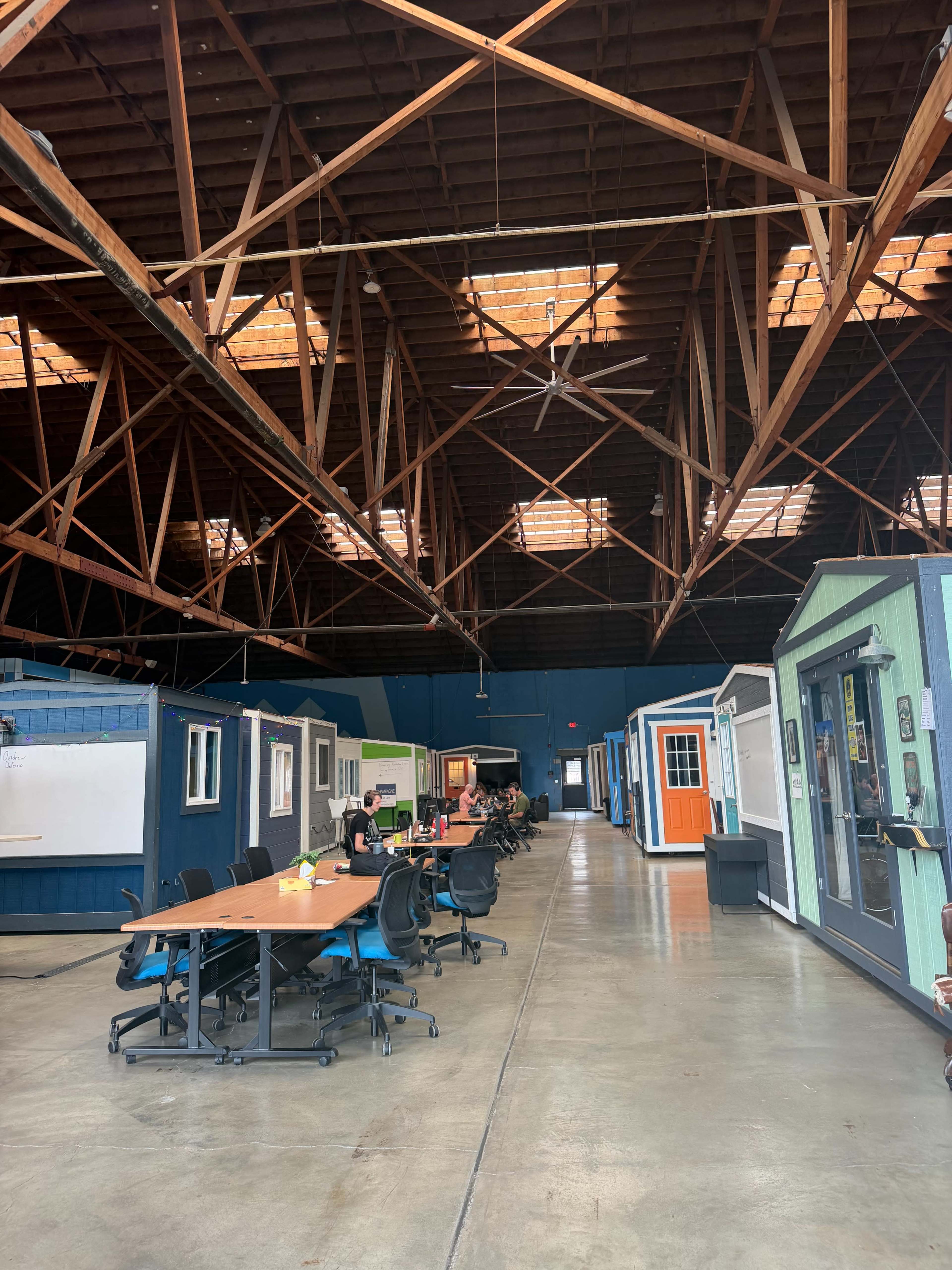 The image shows a spacious indoor area with multiple small, colorful structures resembling cabins or offices, surrounded by tables and chairs for collaboration.
