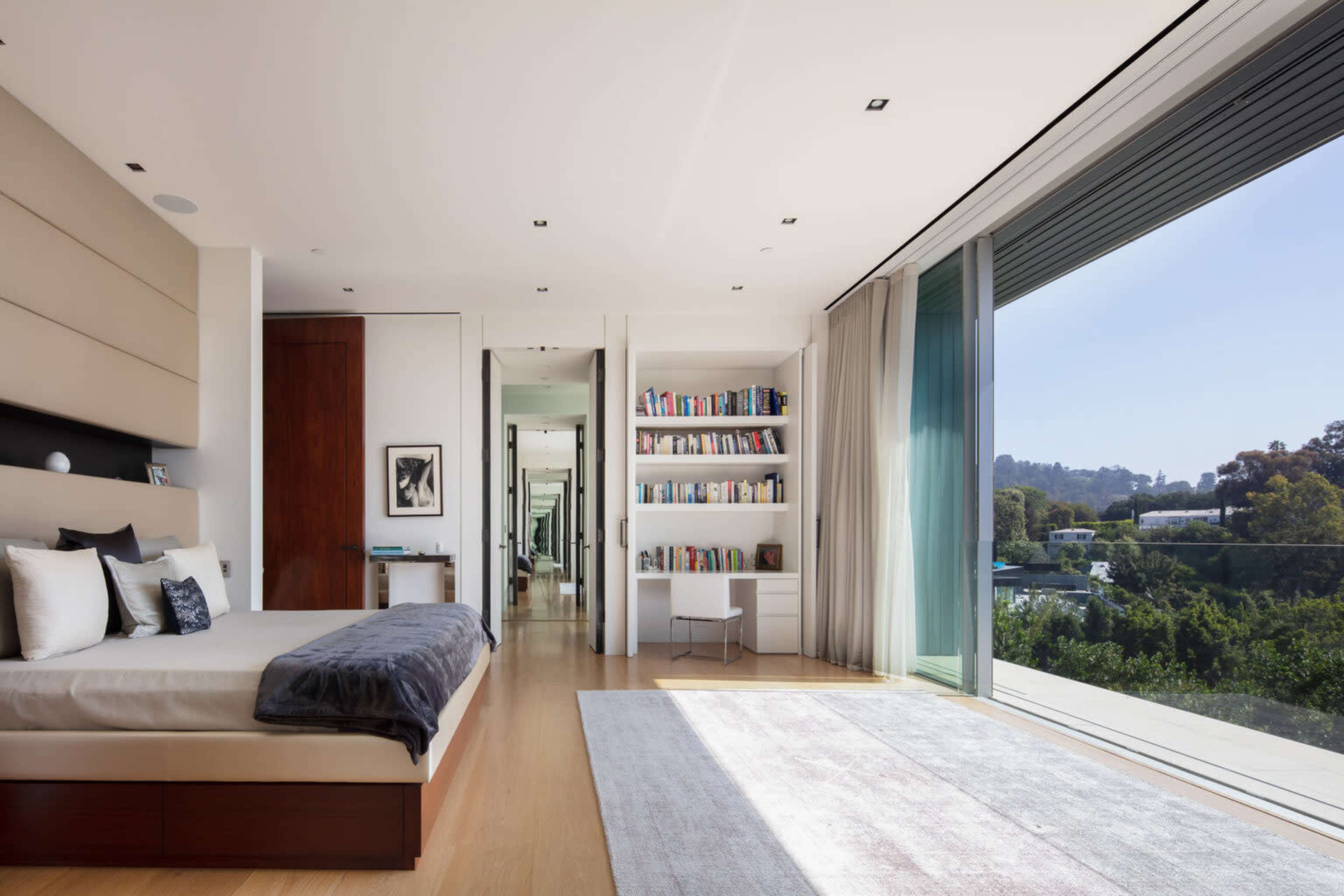 A modern bedroom features a large bed, a bookshelf, and floor-to-ceiling windows overlooking greenery.