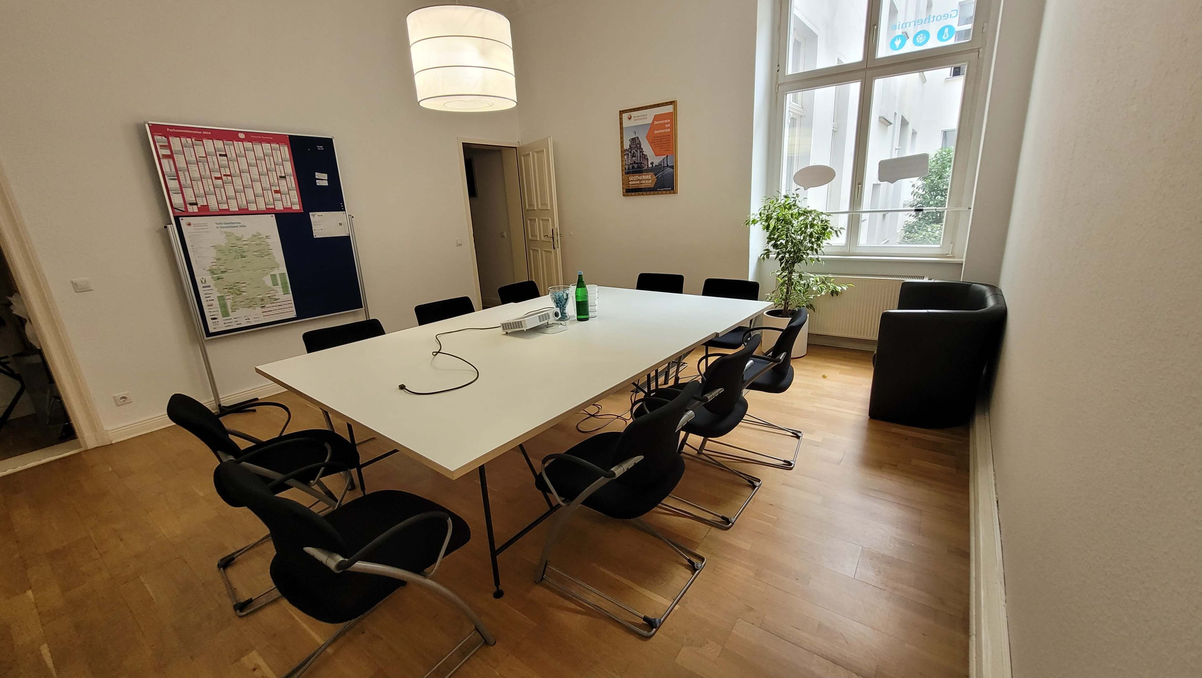 A conference room features a large white table surrounded by black chairs, with a projector on the table and a plant in the corner.