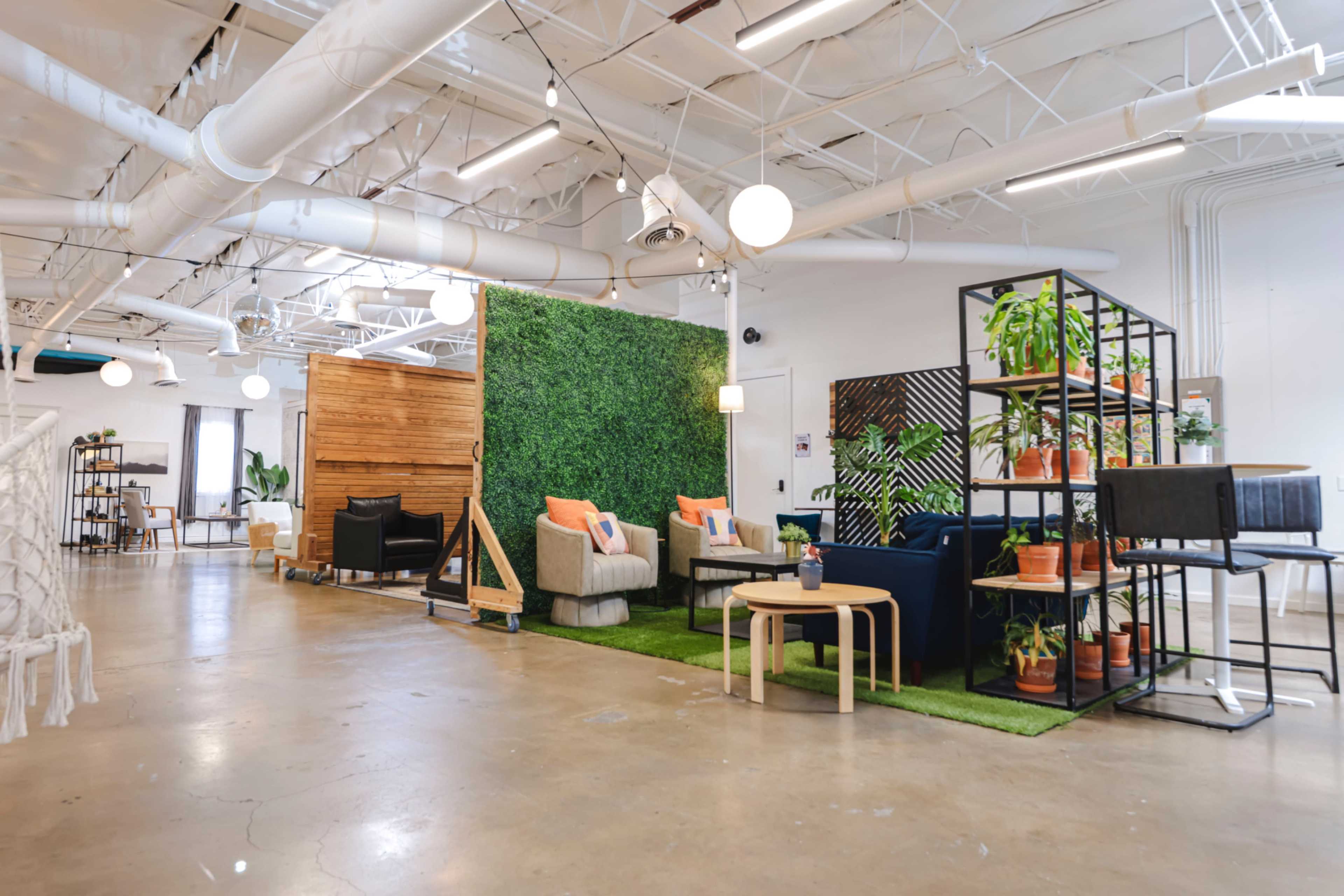 The image shows a modern co-working space featuring various seating arrangements, greenery, and decorative elements in an open layout.