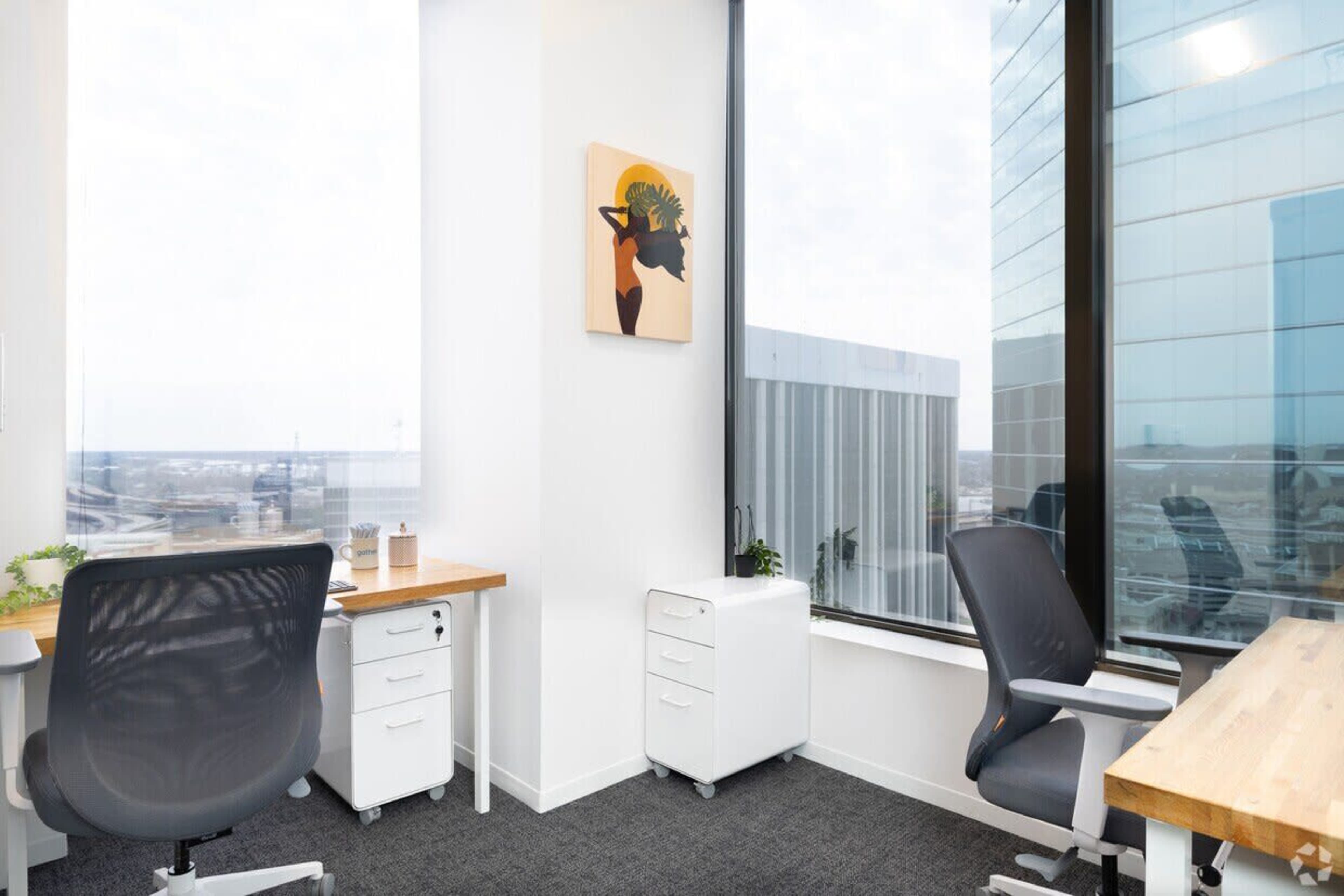 The image shows a modern office space with two desks, a piece of artwork on the wall, and large windows offering a view of the city.