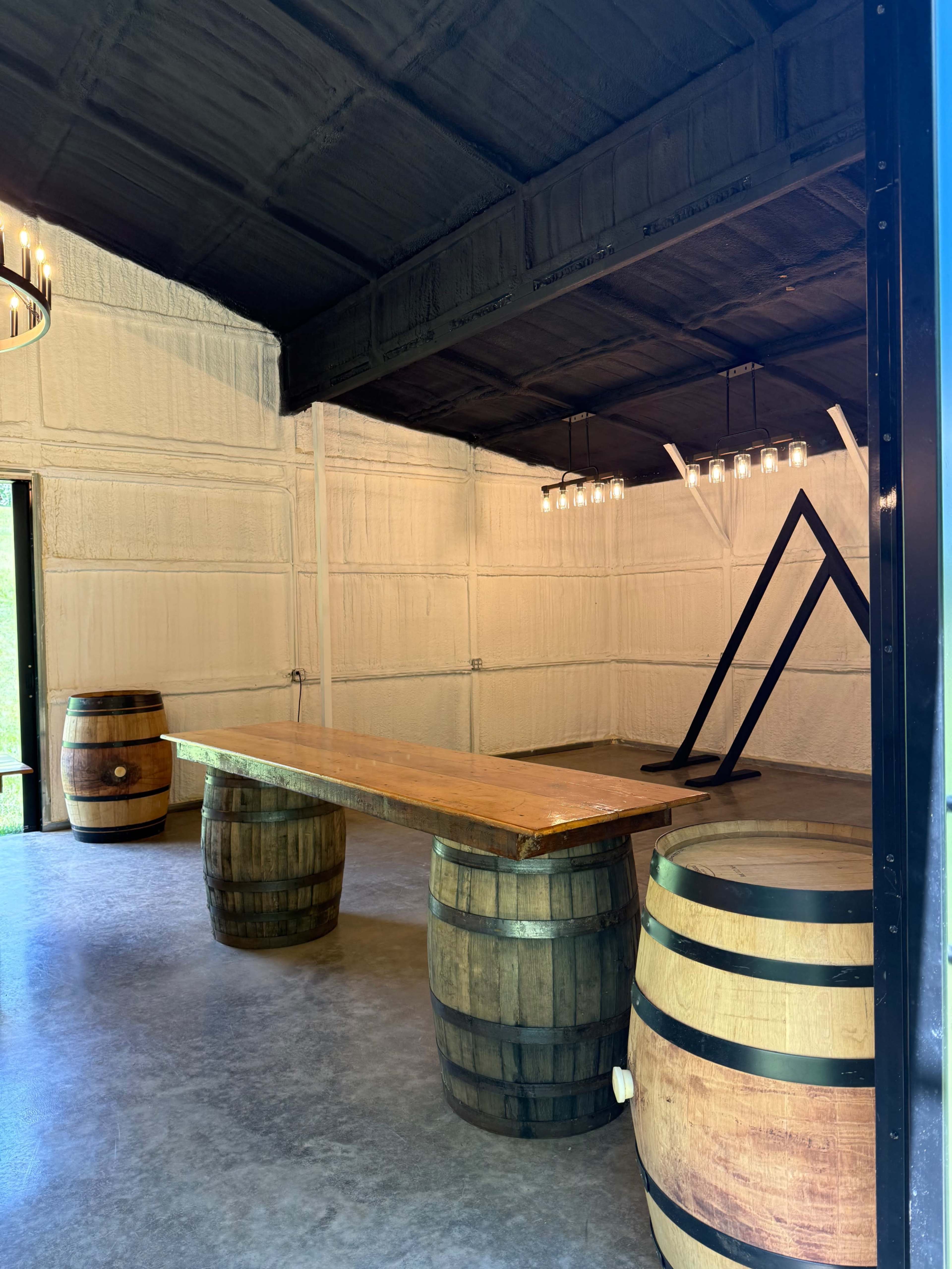 The image shows a rustic room with a wooden table supported by two barrels and a triangular structure in the background, all under a high ceiling with pendant lights.