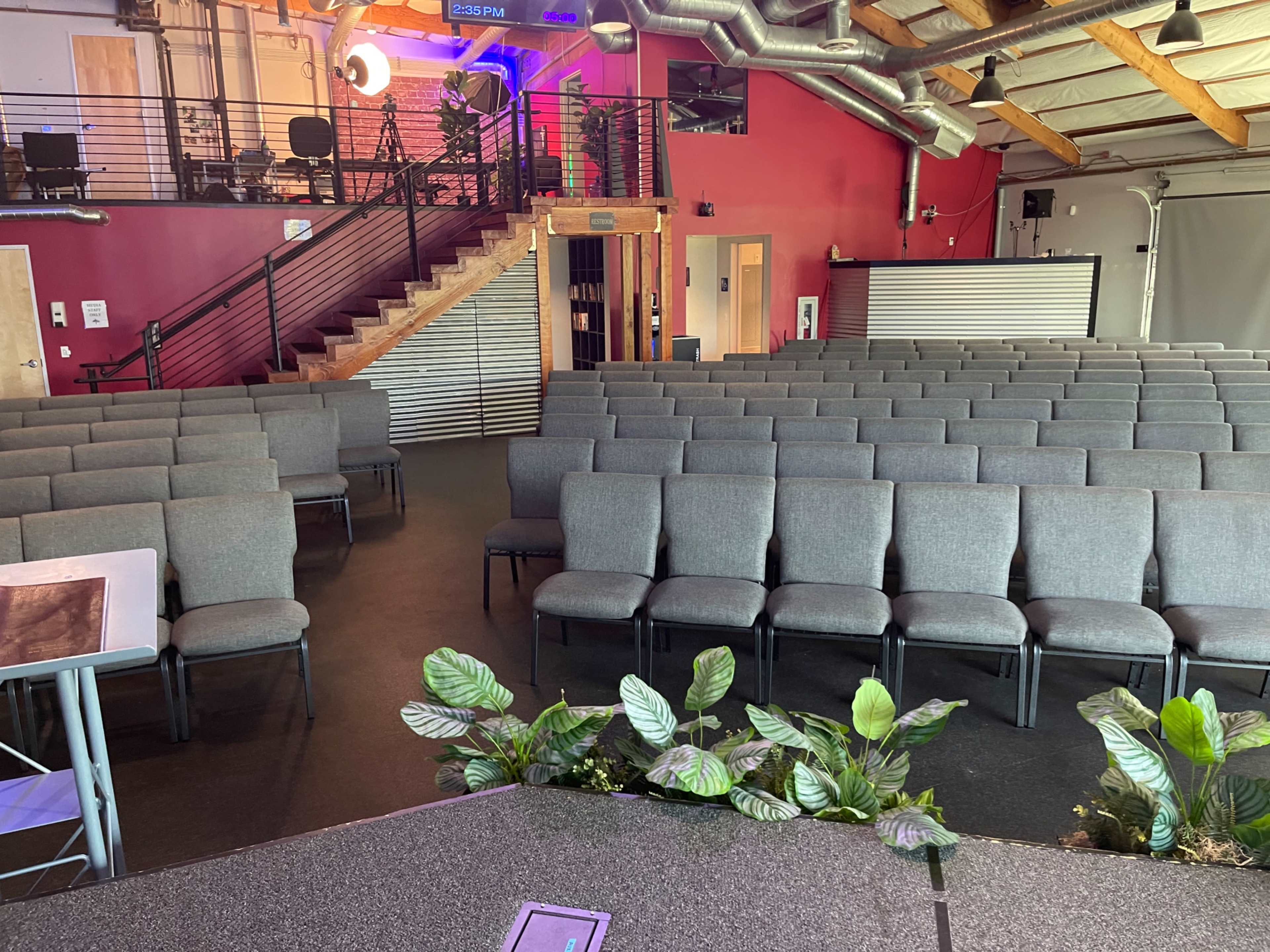 The image shows a spacious auditorium filled with neatly arranged gray chairs, facing a presentation stage adorned with potted plants.