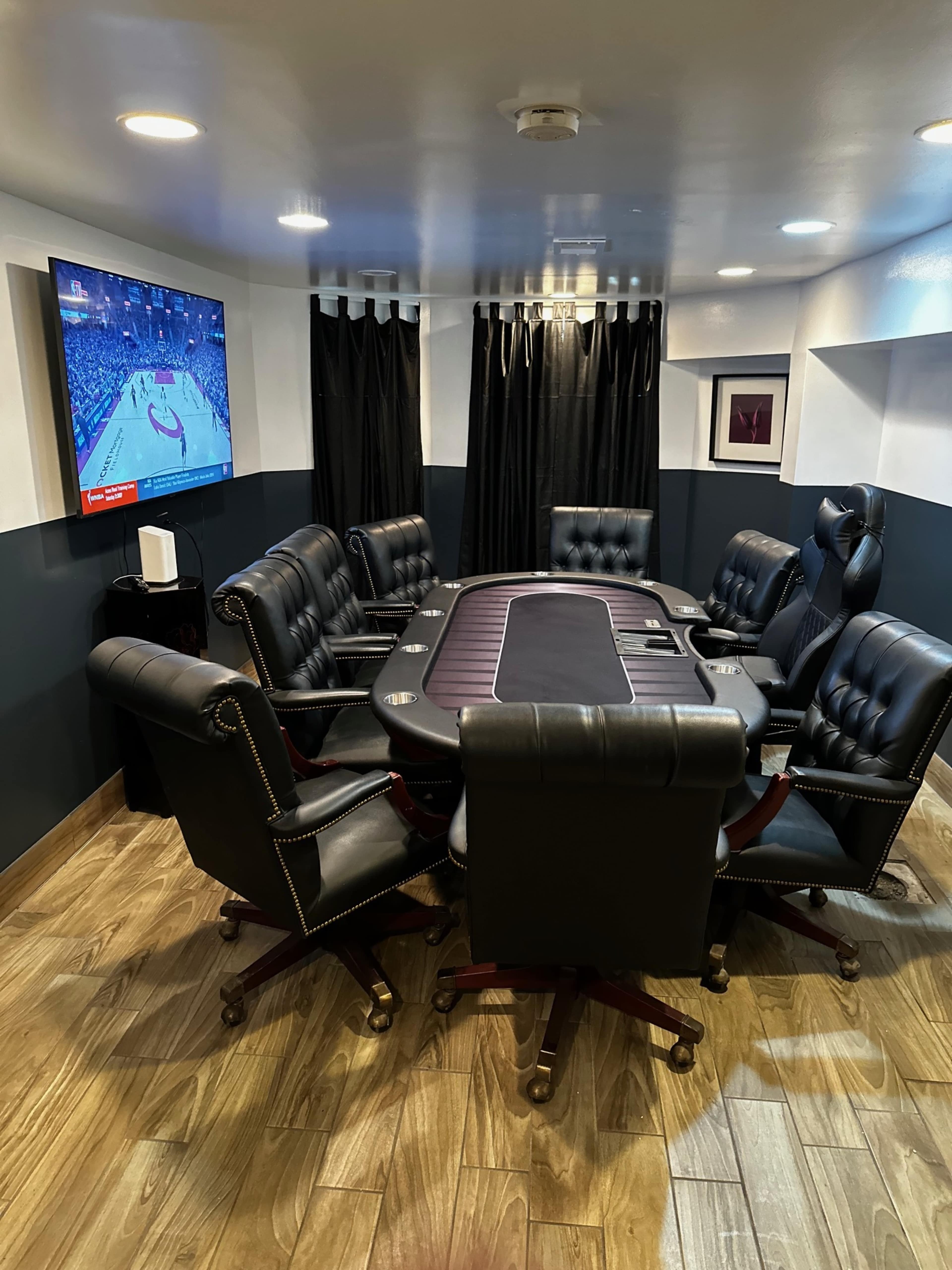 A poker room features a central table surrounded by eight black leather chairs and a television showing a sports game on the wall.