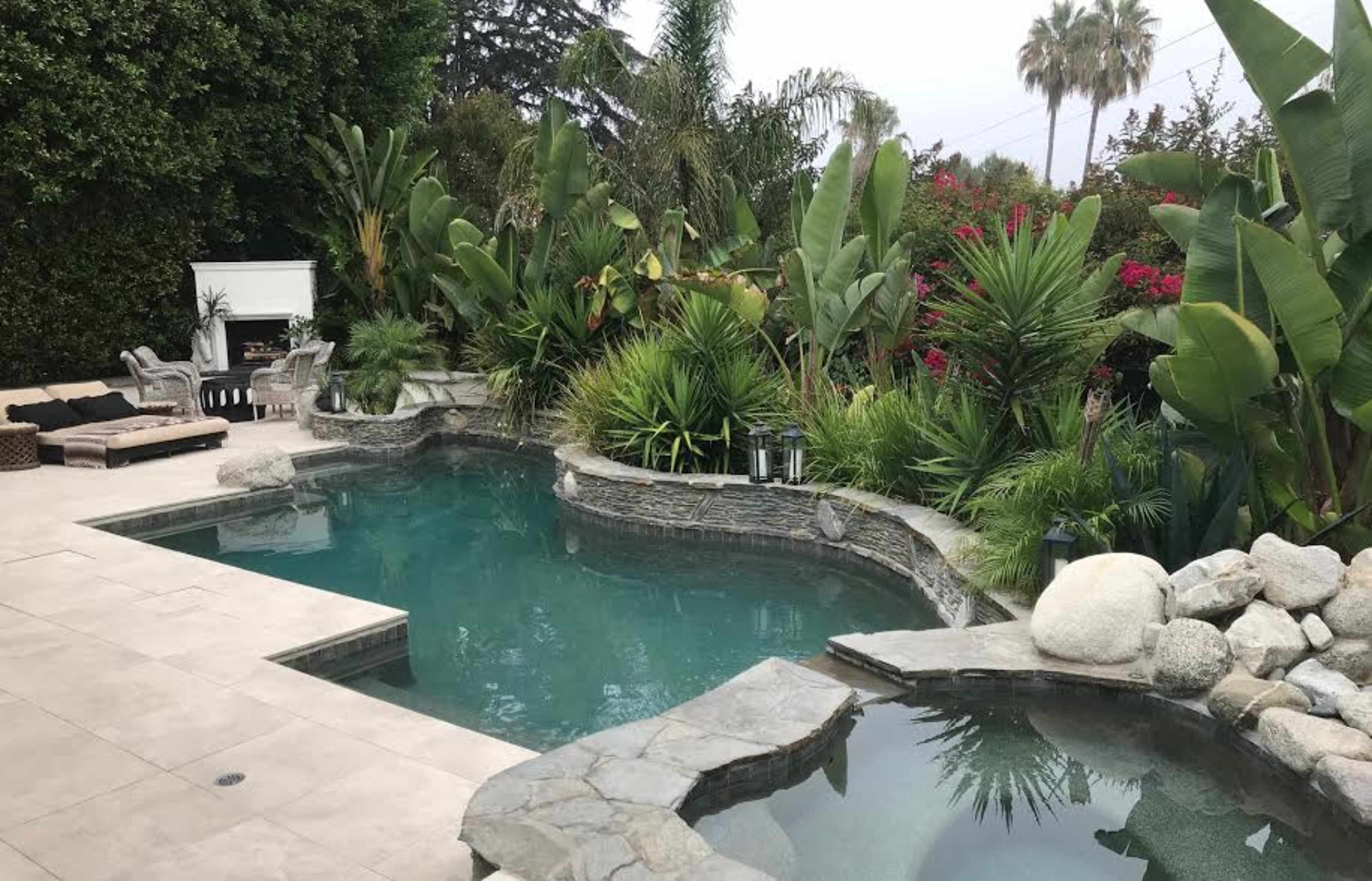 A landscaped backyard featuring a swimming pool surrounded by tropical plants and a stone patio with lounge chairs.