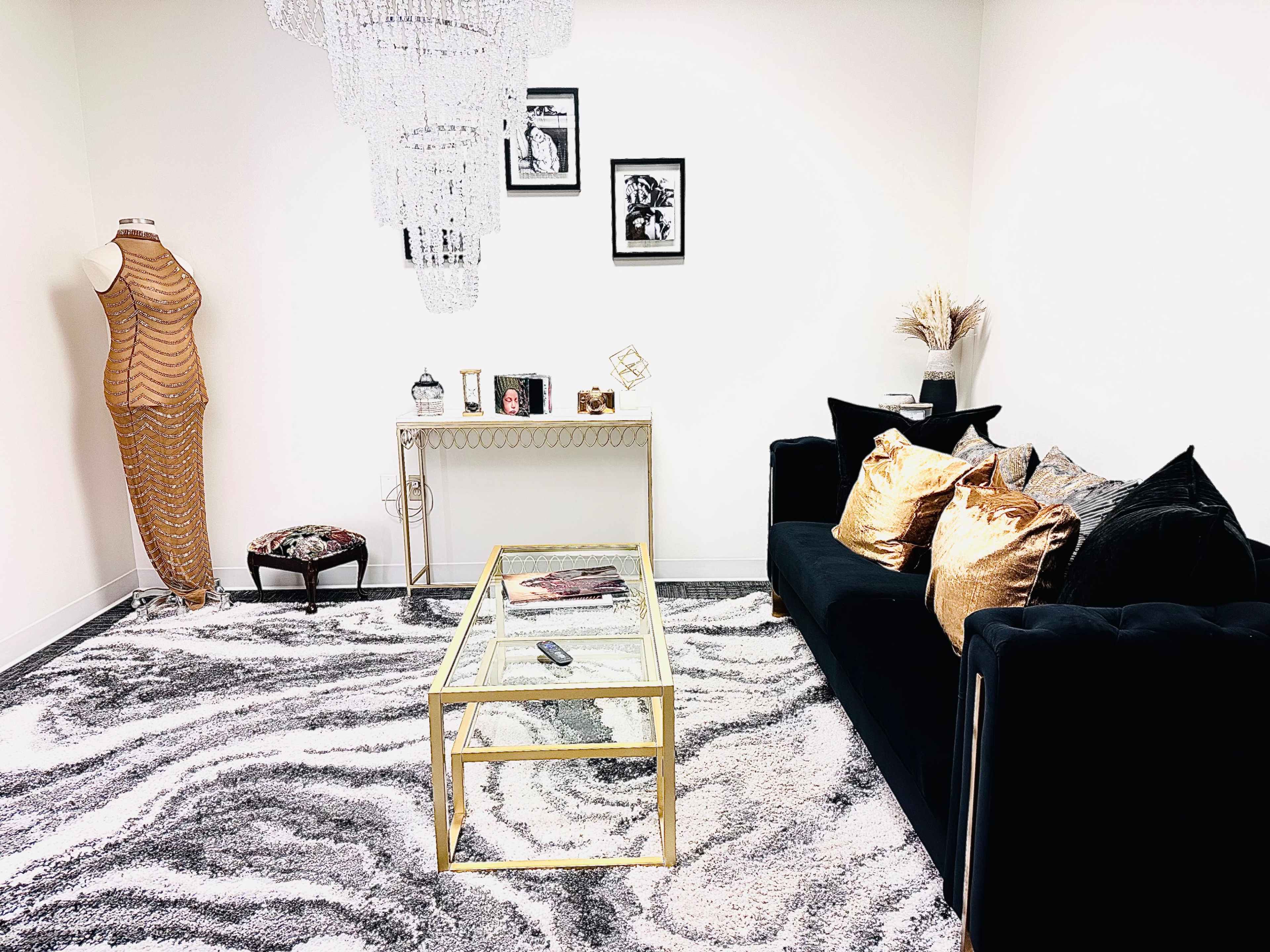 A stylish interior featuring a black sofa with decorative cushions, a gold-framed glass coffee table, a dress form, and a chandelier, all set on a patterned rug.