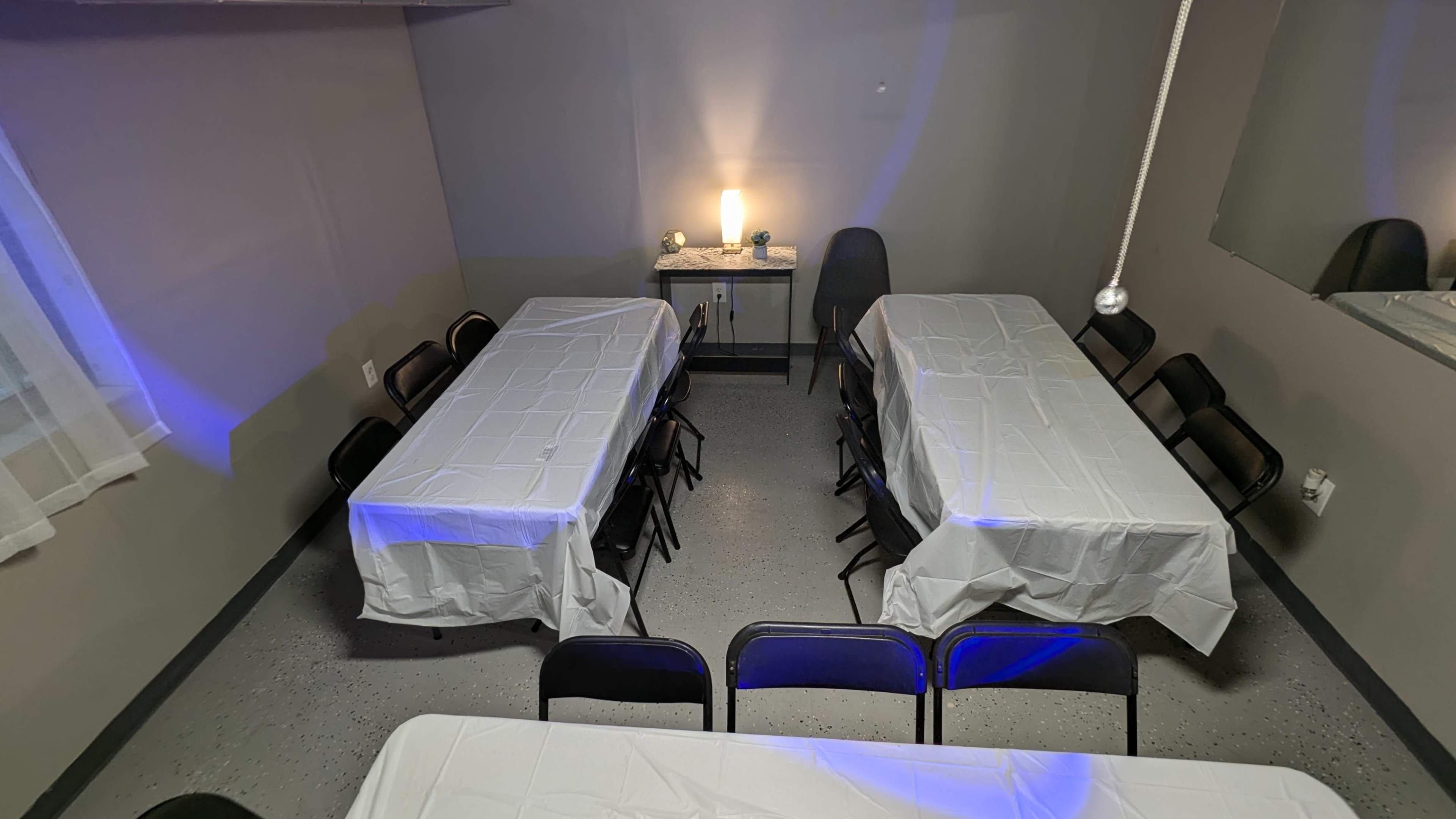 The image shows a small room set up with two rectangular tables covered in white tablecloths and surrounded by black chairs, with a lamp on a side table illuminating the space.