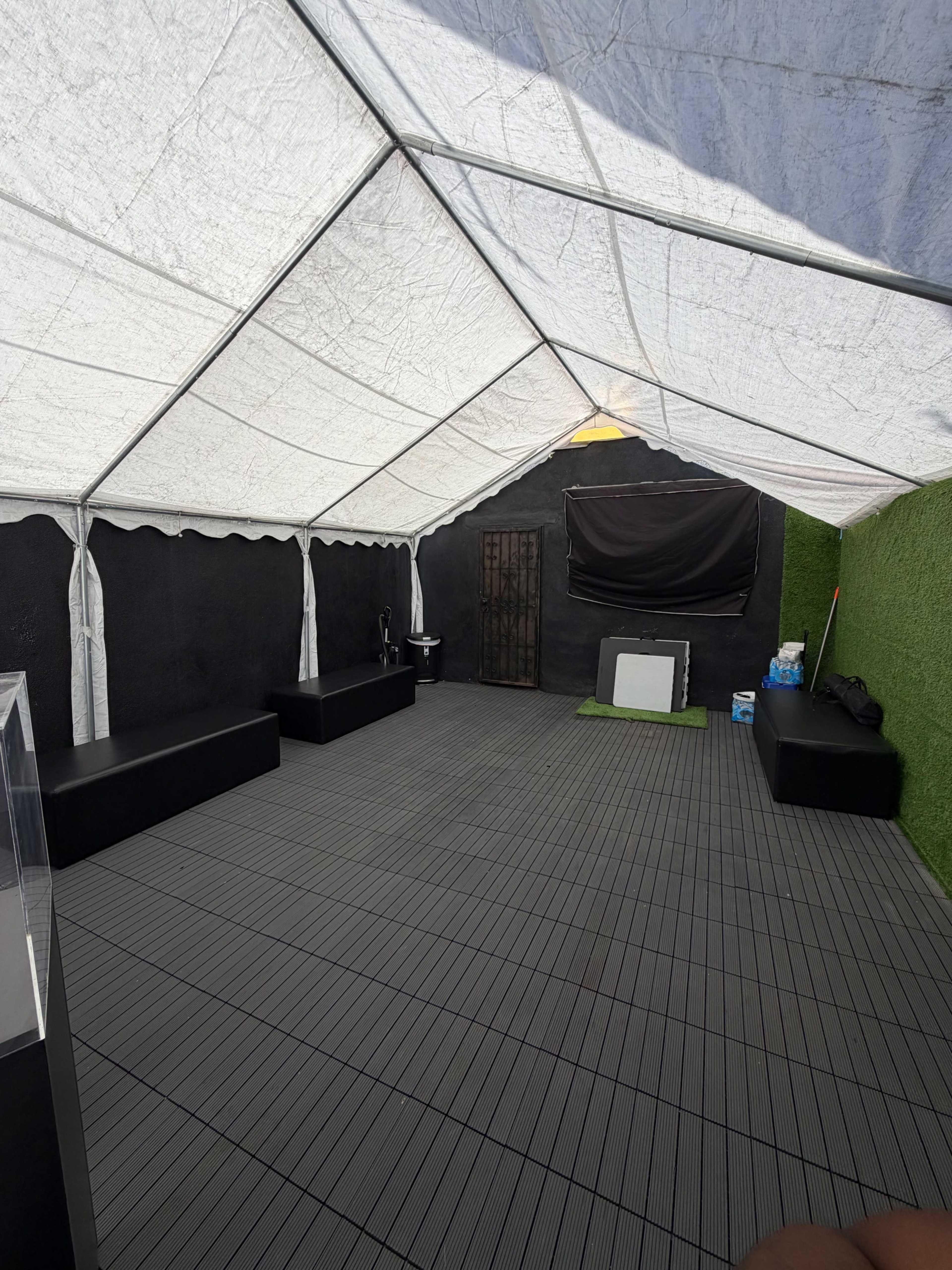 The image shows the interior of a tent with gray flooring, three black benches, and a black backdrop, set against a green artificial grass wall.