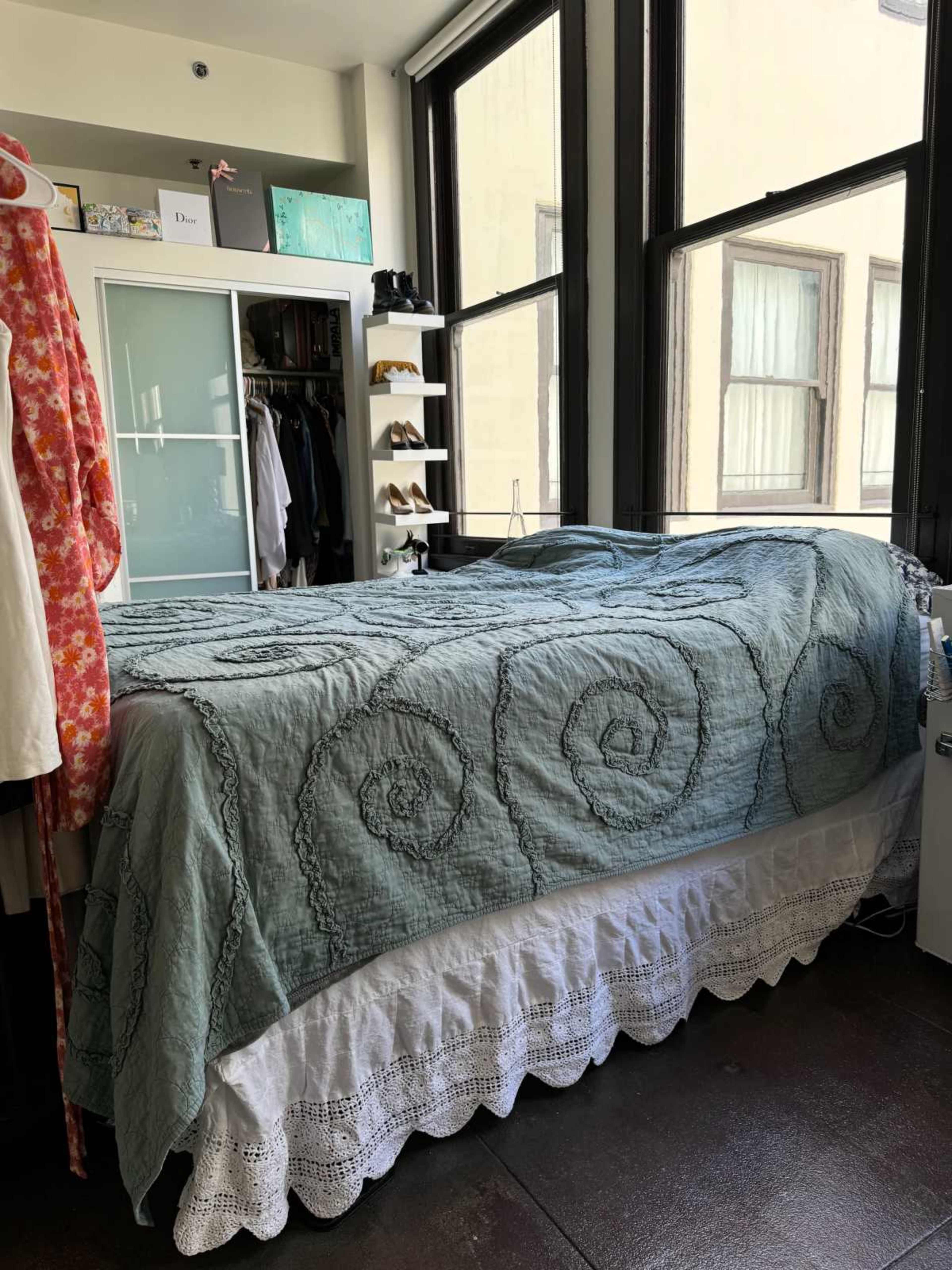The image shows a bedroom with a bed covered by a quilt, a wardrobe with clothes, and large windows allowing natural light to enter.