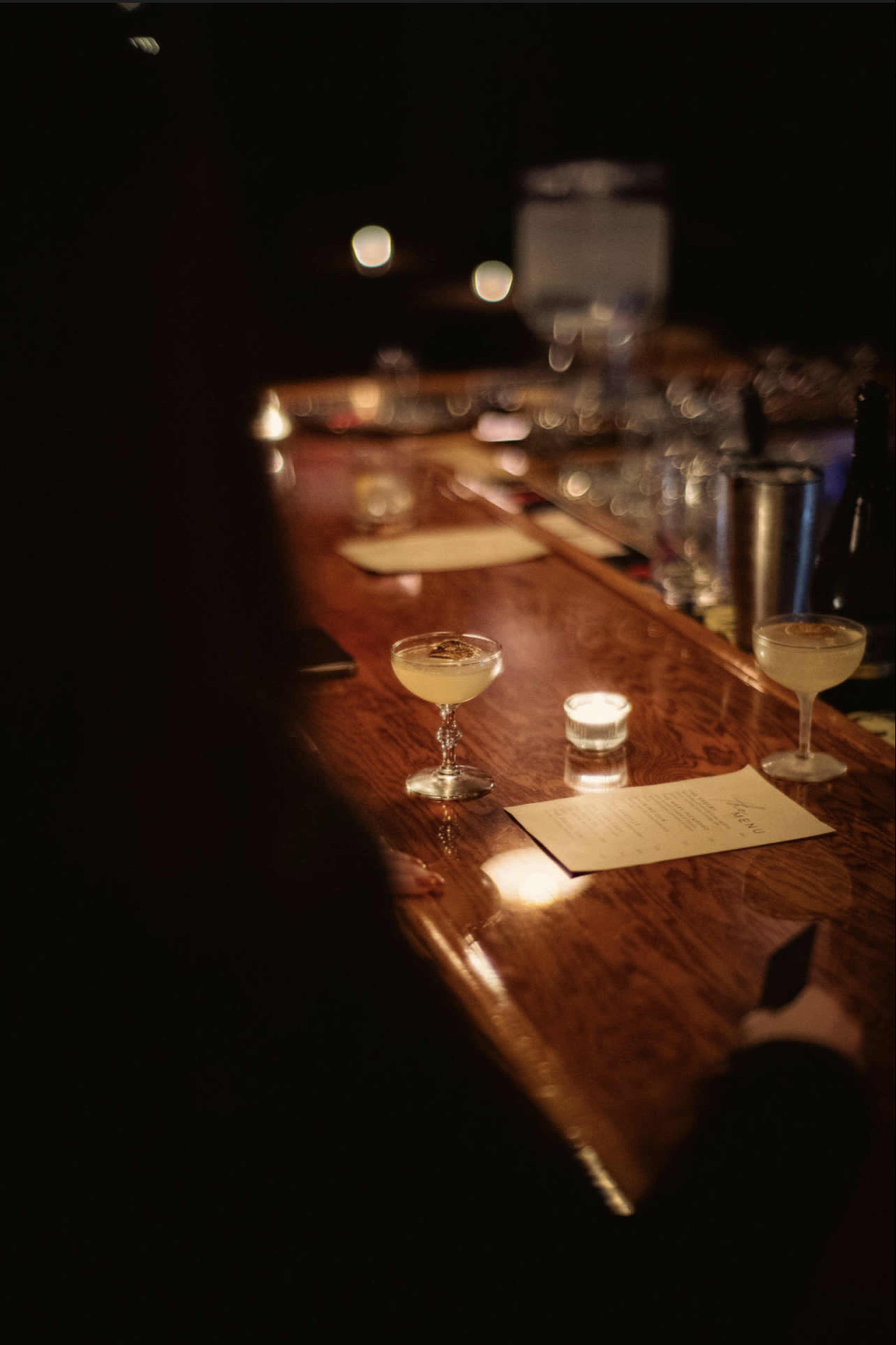 A dimly lit bar is depicted, featuring a long wooden counter with a cocktail glass and a candle placed on top, alongside a menu.