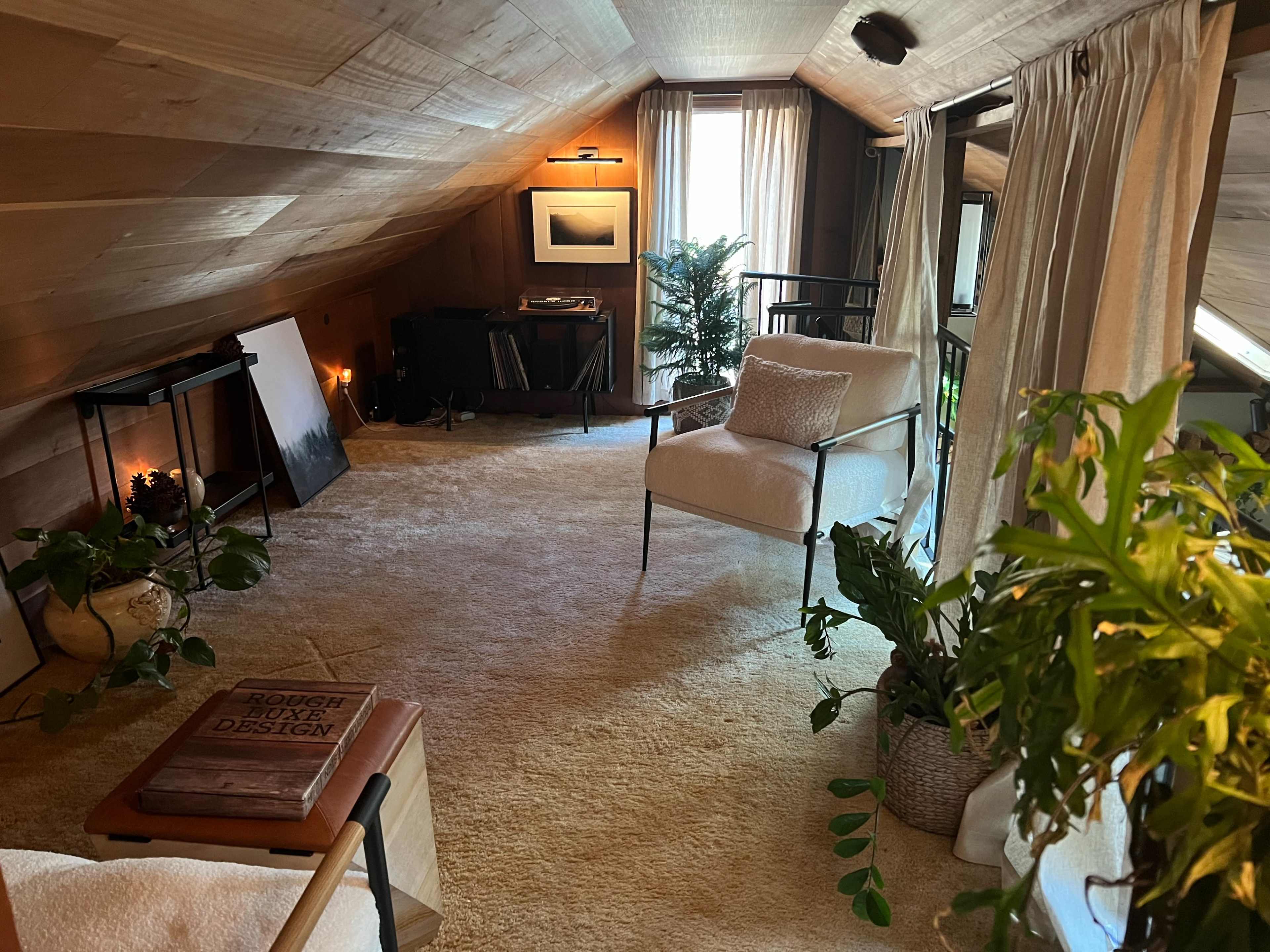 The image shows a cozy attic room with wooden walls, a sloped ceiling, a white armchair, plants, and various decorative elements.