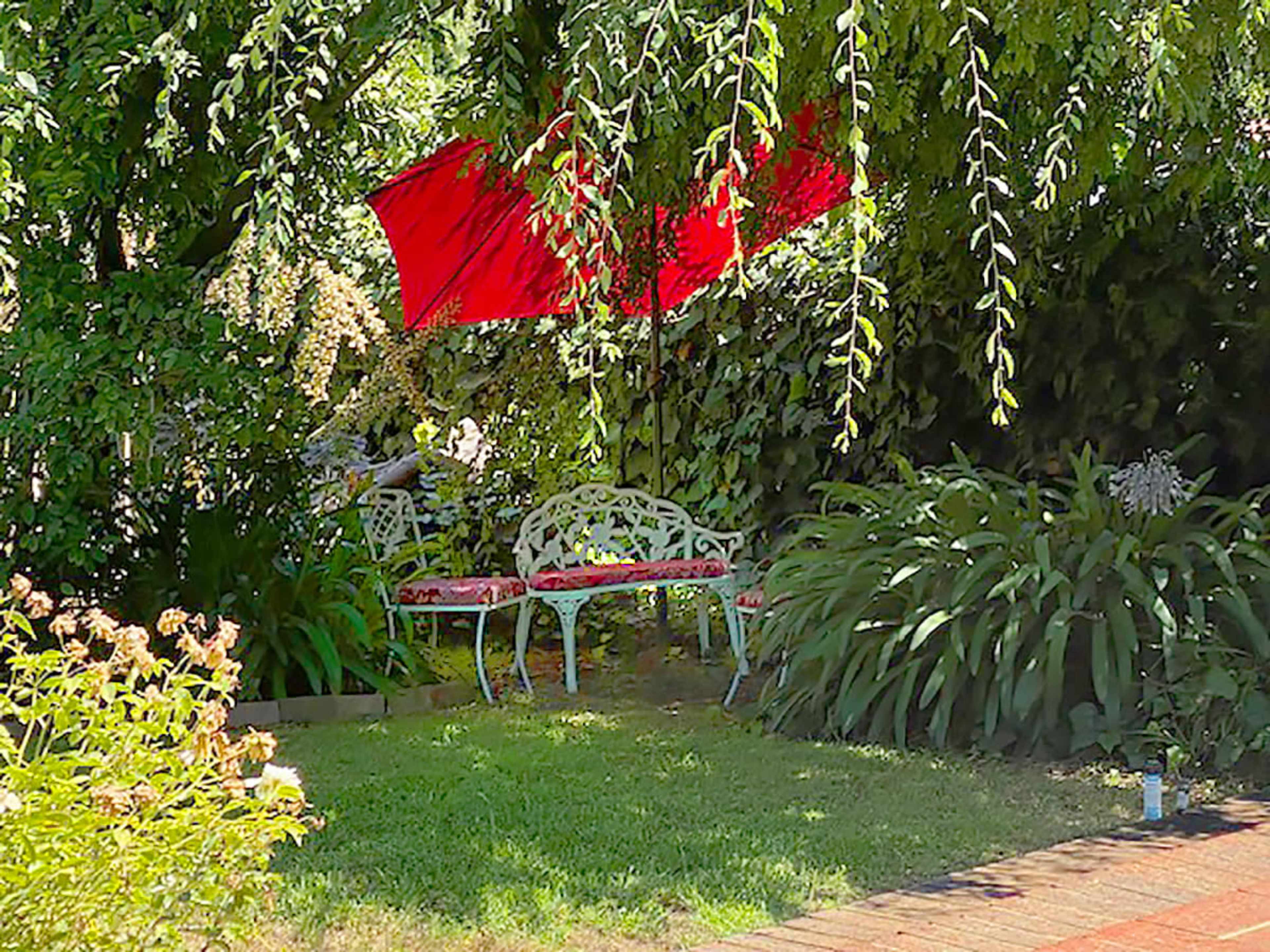 A red umbrella shades two white metal benches surrounded by lush green plants in a garden setting.