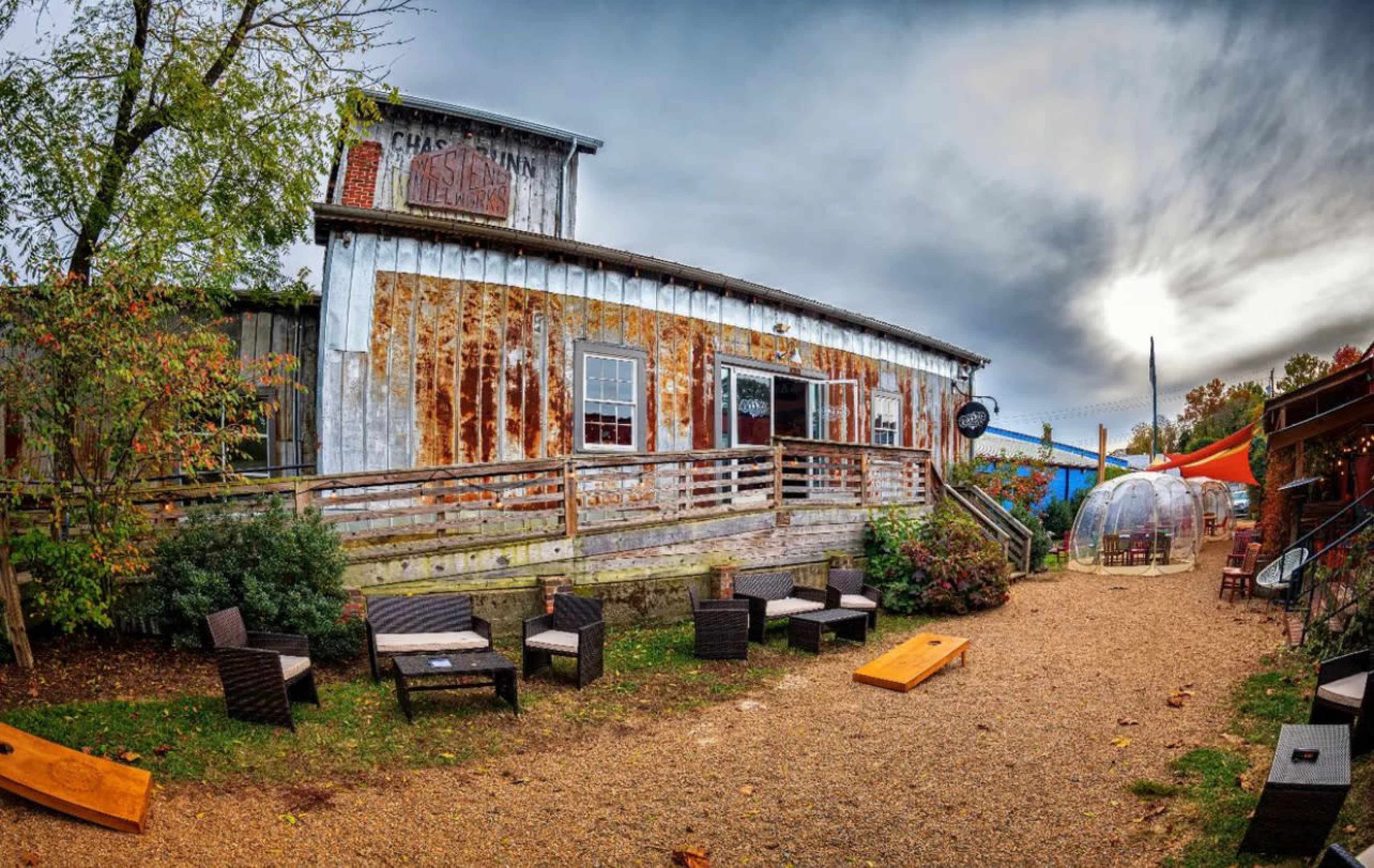 A rustic building with weathered wood siding, surrounded by a gravel path and outdoor seating areas, along with colorful canopies in the background.