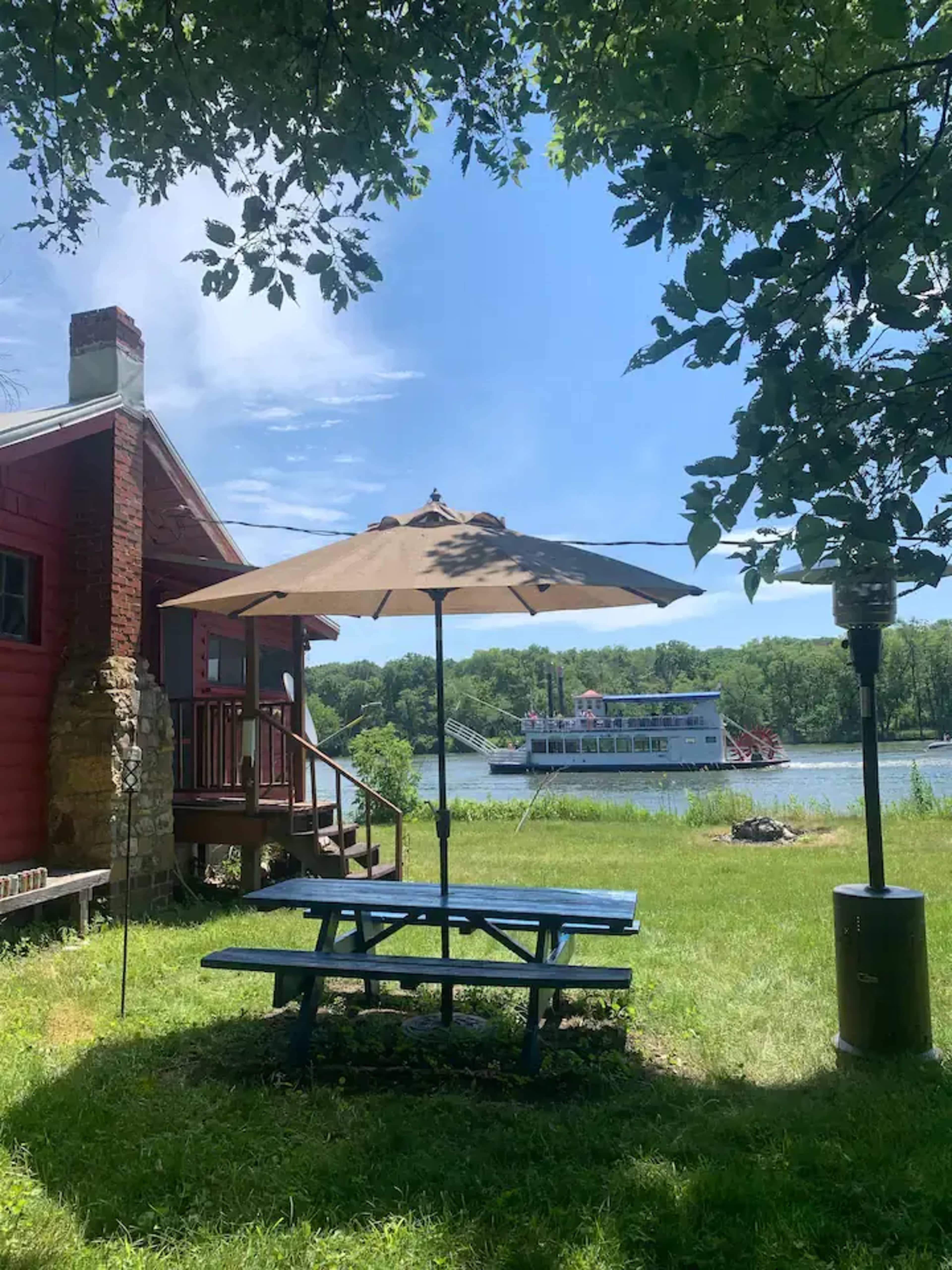 A picnic table with an umbrella is set in a grassy area next to a river, with a red building nearby and a boat in the background.