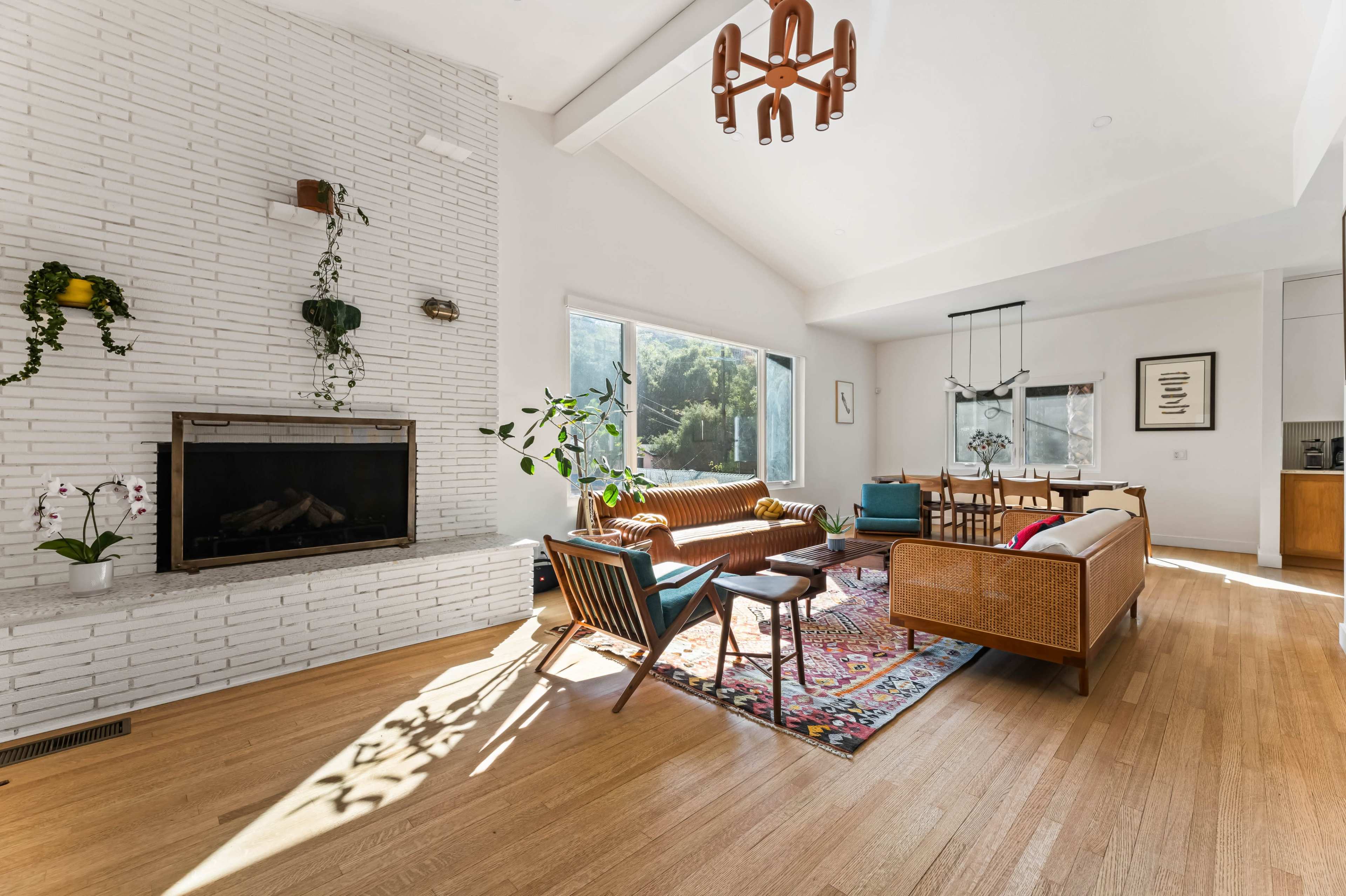 The image shows a bright, modern living room with a fireplace, large windows, and a mixture of seating options arranged around a colorful area rug.