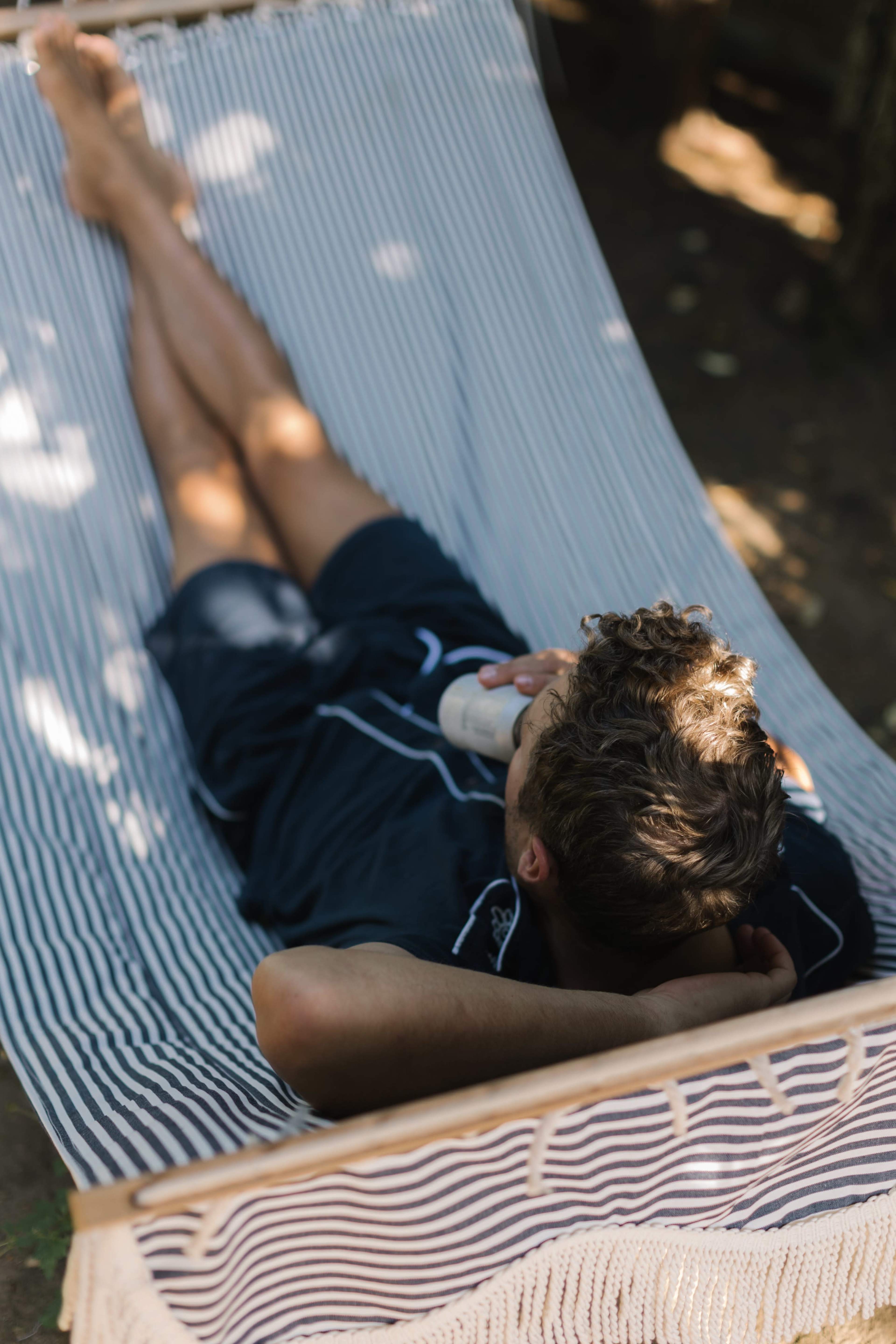 A person relaxes in a hammock, holding a drink while reclining with their feet stretched out.