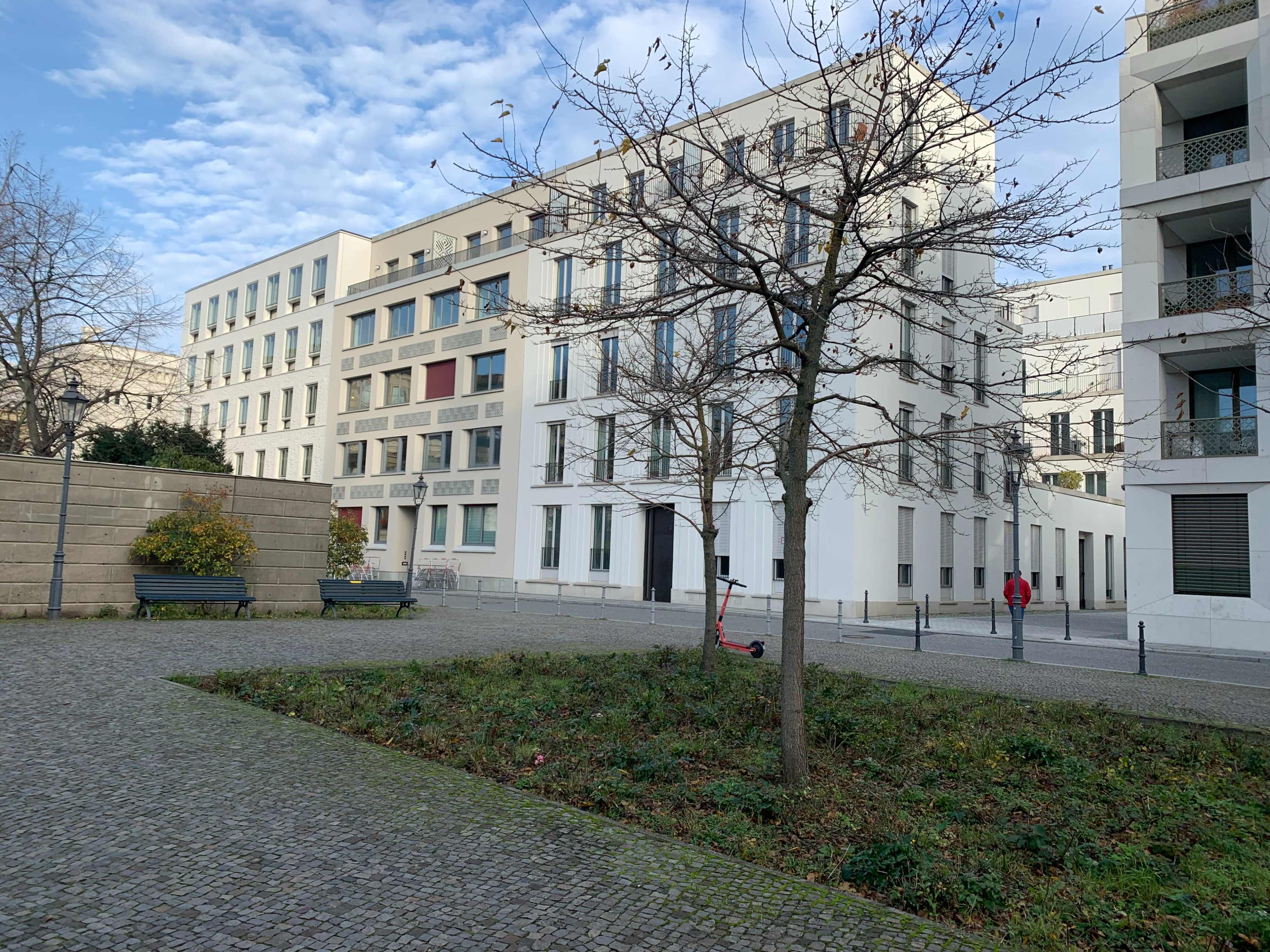 A sparse urban plaza features a tree and two benches, surrounded by modern white buildings under a cloudy sky.