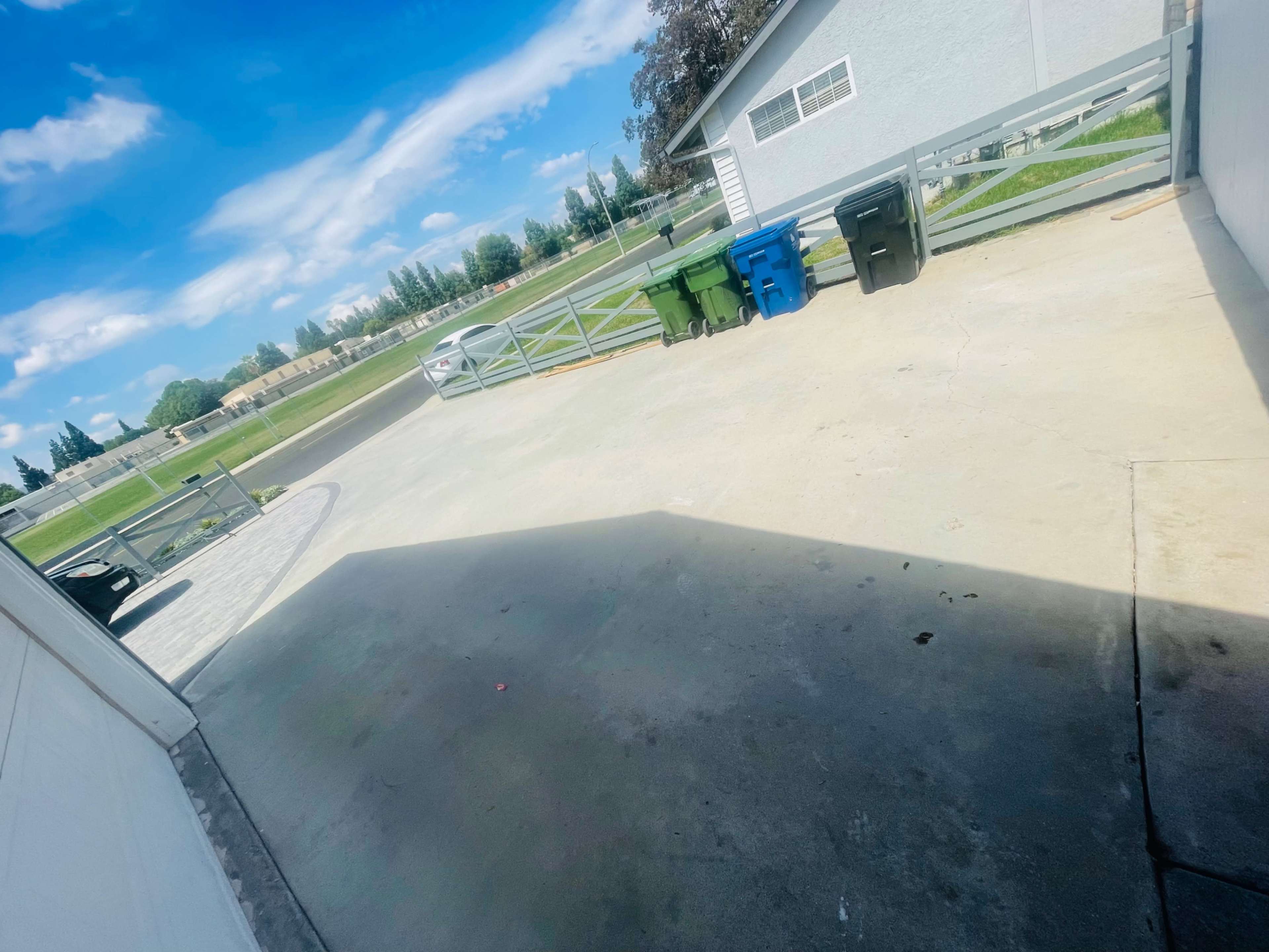 A concrete driveway with three trash bins lined up against a fence and a grassy area in the background.