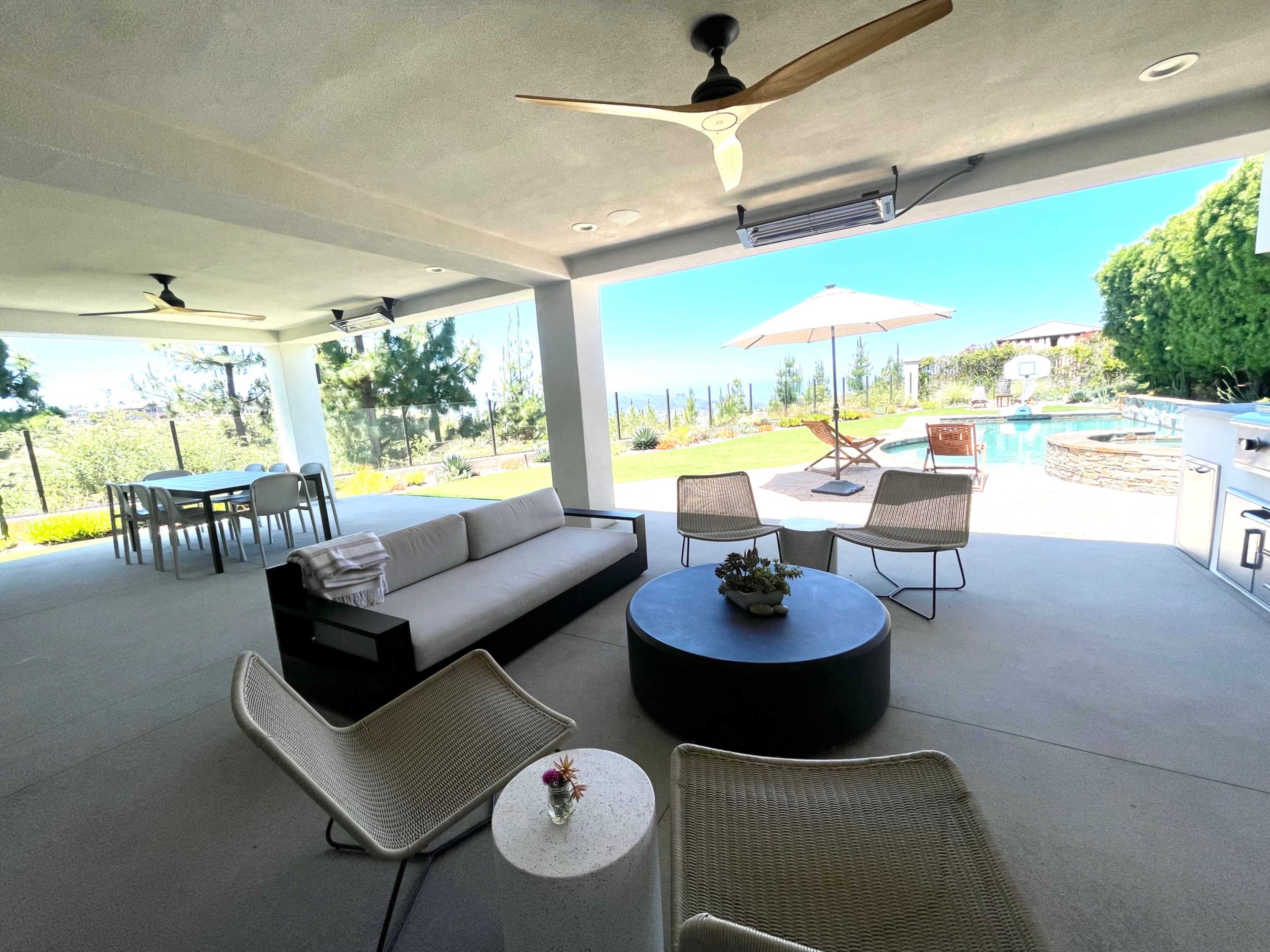 A covered outdoor living area featuring a sectional sofa, two chairs, a round coffee table, and a dining table, with a swimming pool and lush greenery visible in the background.
