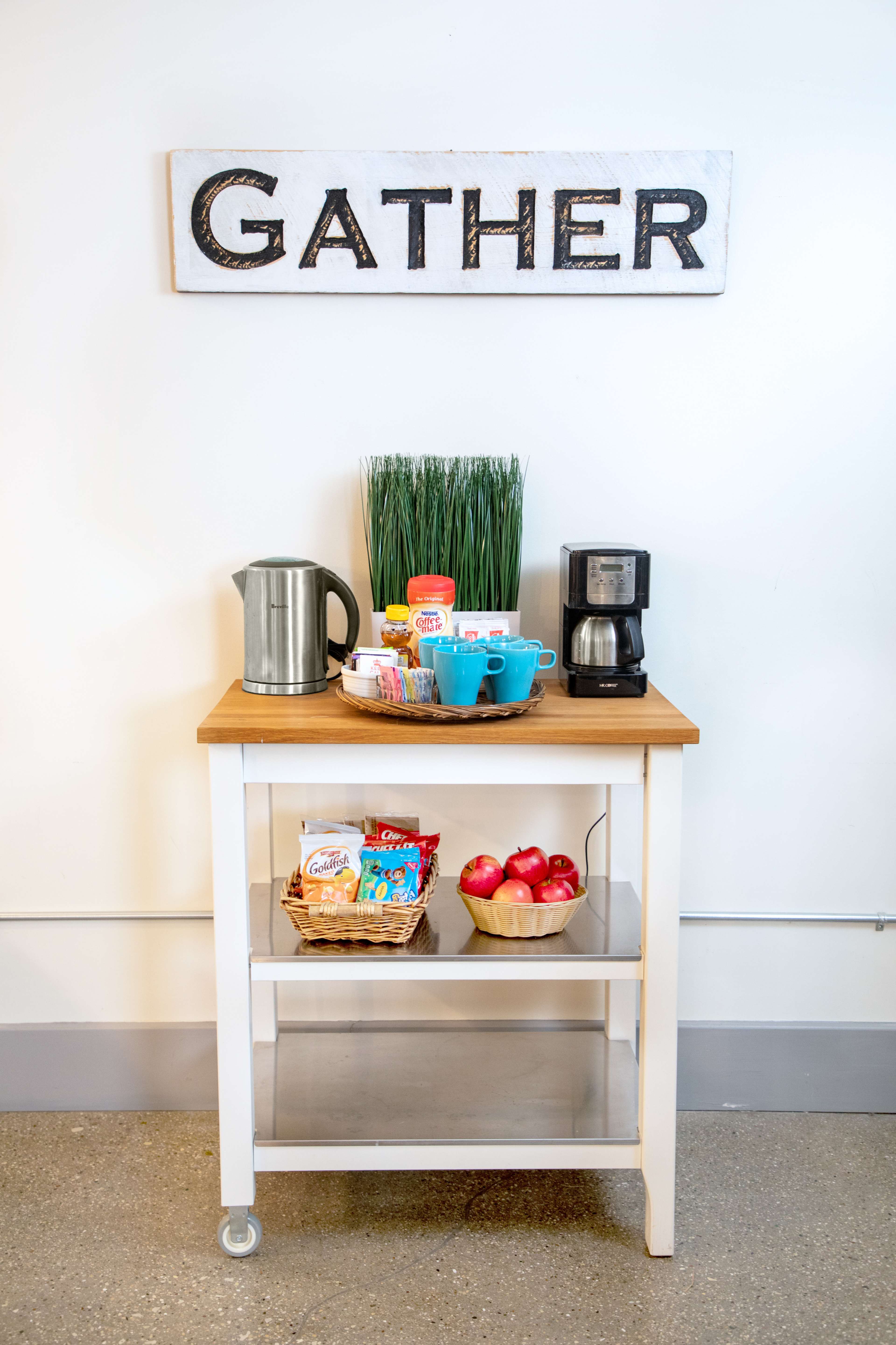 A wooden table with a top shelf featuring a kettle, coffee maker, cups, and a variety of snacks, and a bottom shelf containing a basket of apples and other items, all beneath a "GATHER" sign.