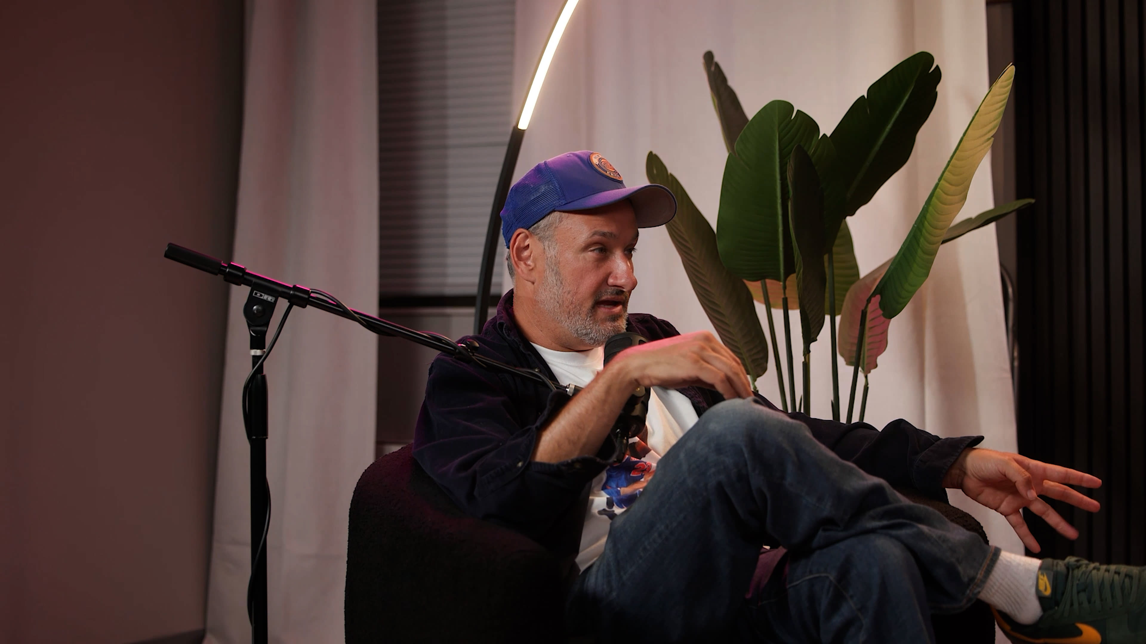 A man wearing a blue cap sits in a black chair with plants in the background, gesturing while talking.