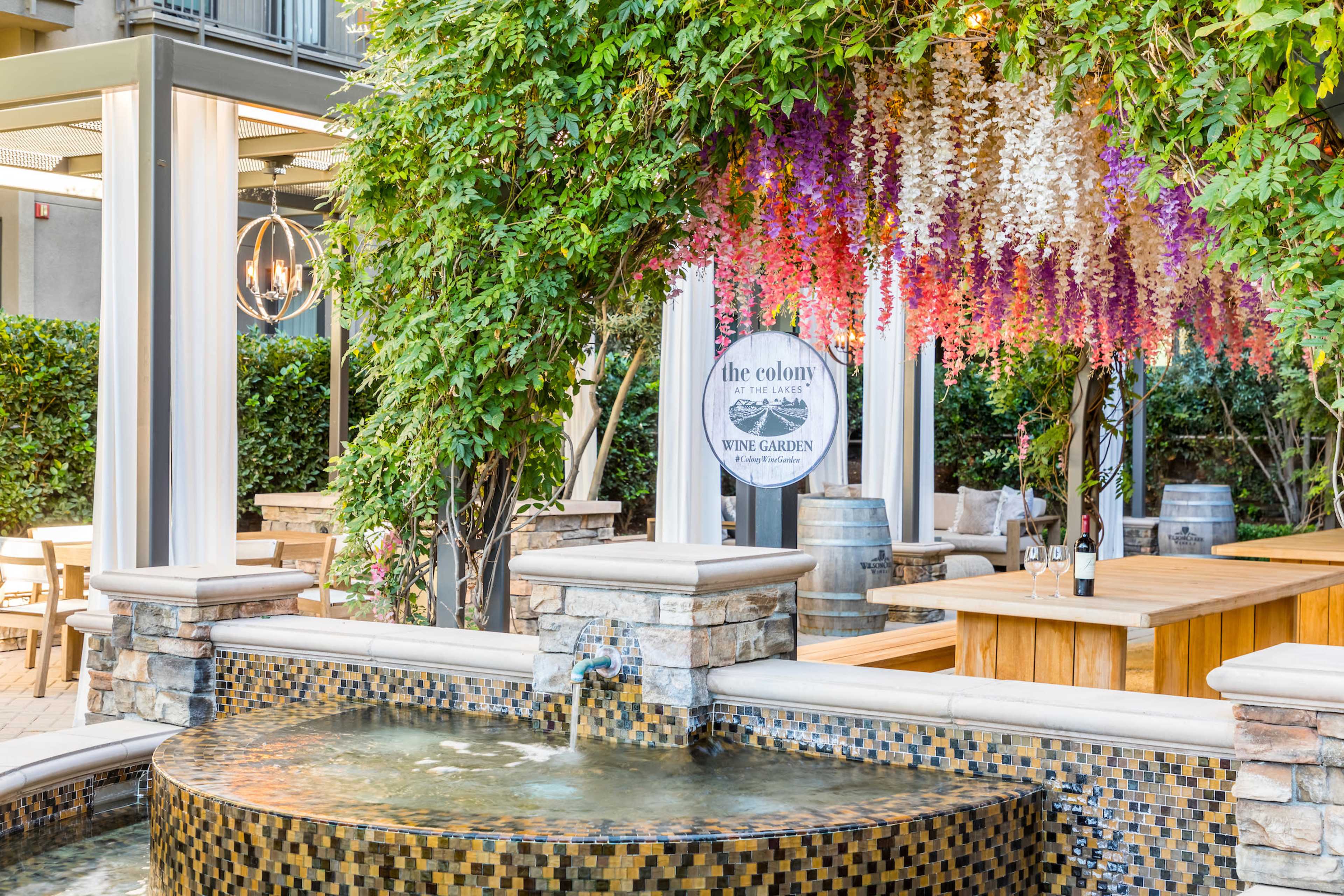 A fountain surrounded by lush greenery and decorative flowers, with outdoor dining furniture visible in the background.
