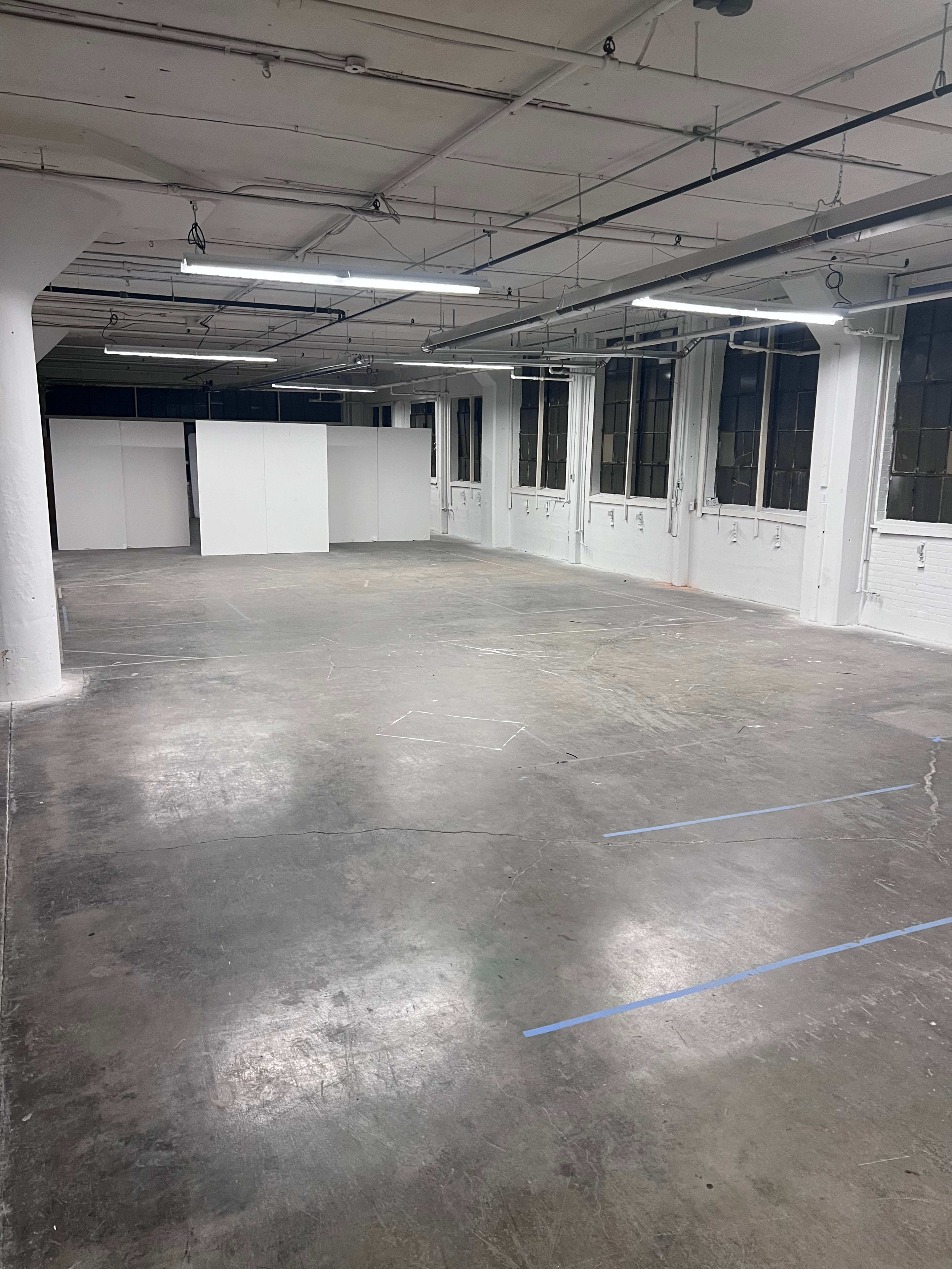 The image shows a large, empty room with concrete flooring, white walls, and a few white partitions in the background.