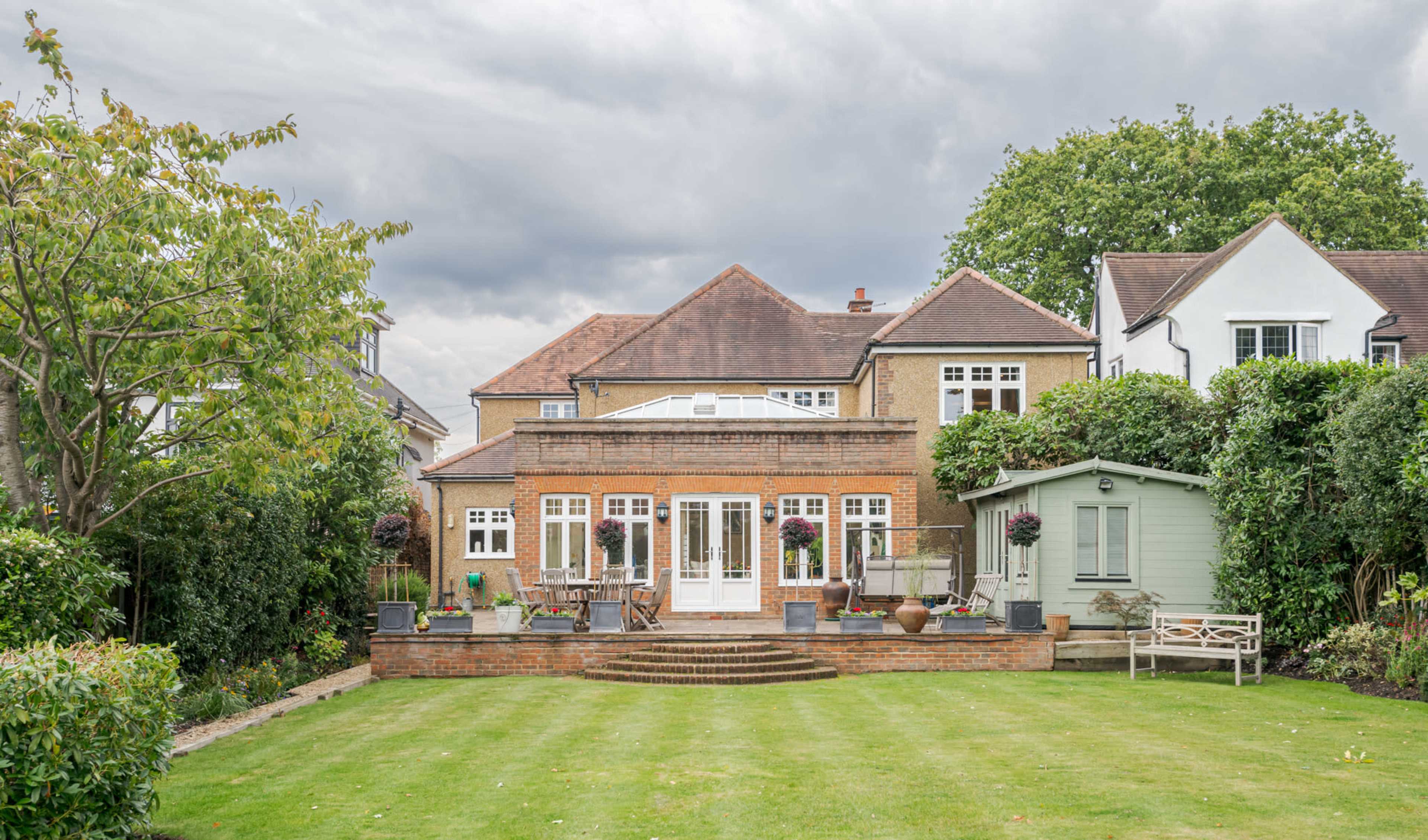A large brick house with a flat-roofed extension is set in a green garden, featuring a patio area and a shed at the rear.