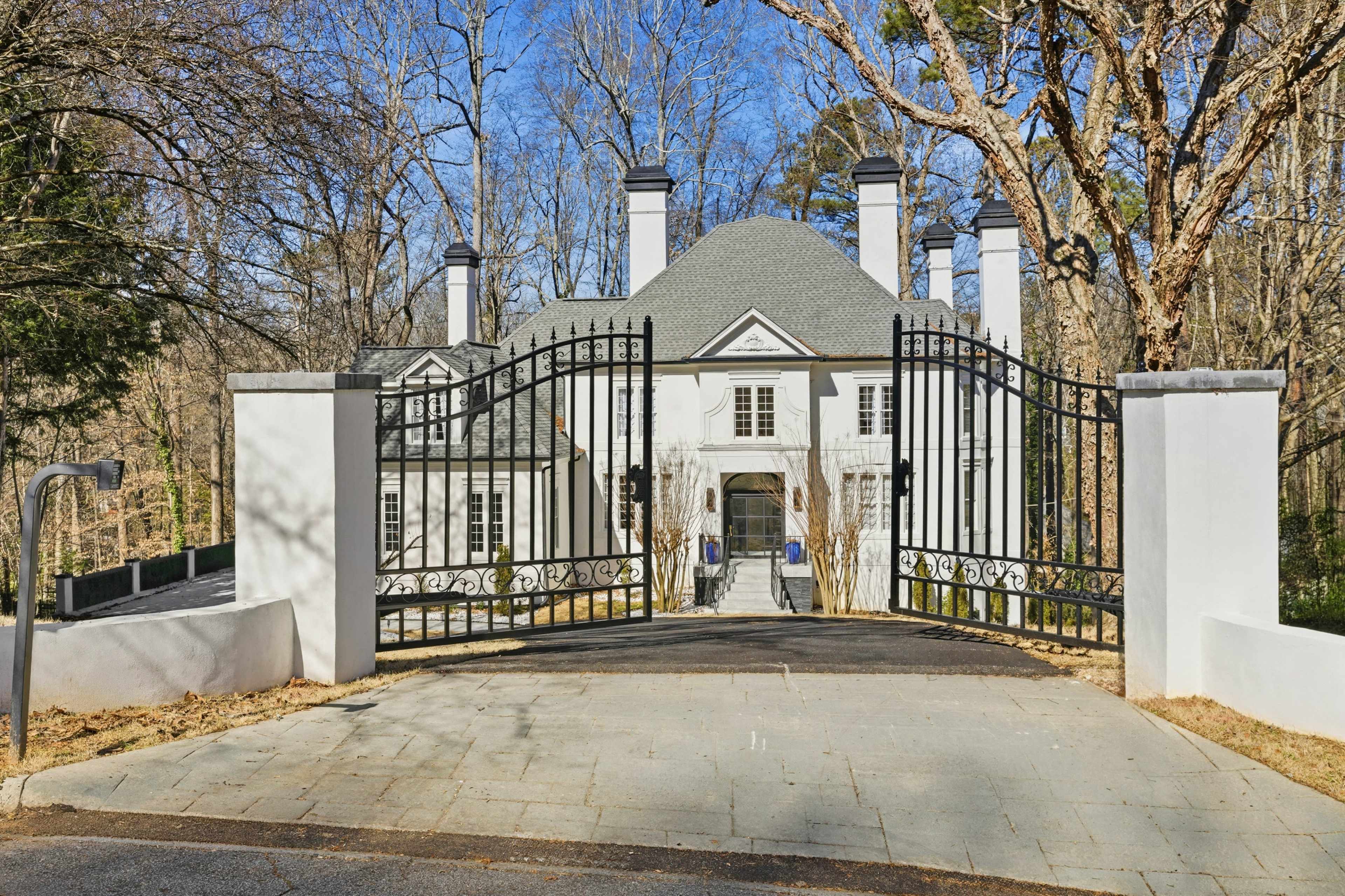 A large, gated mansion with a gray roof and white exterior is surrounded by trees.
