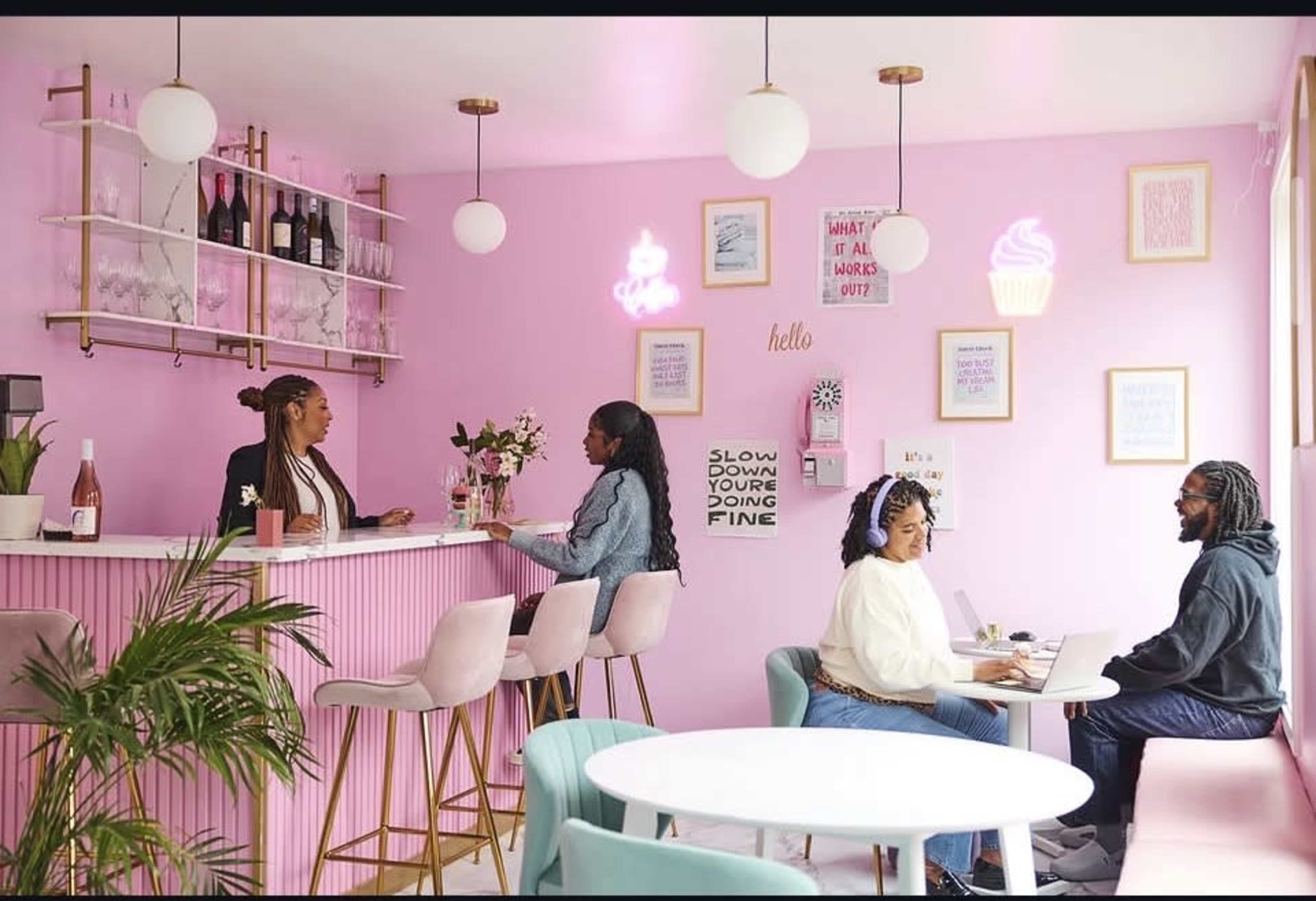 A colorful cafe features a pink interior with customers sitting at a stylish bar and tables, enjoying drinks and working on laptops.