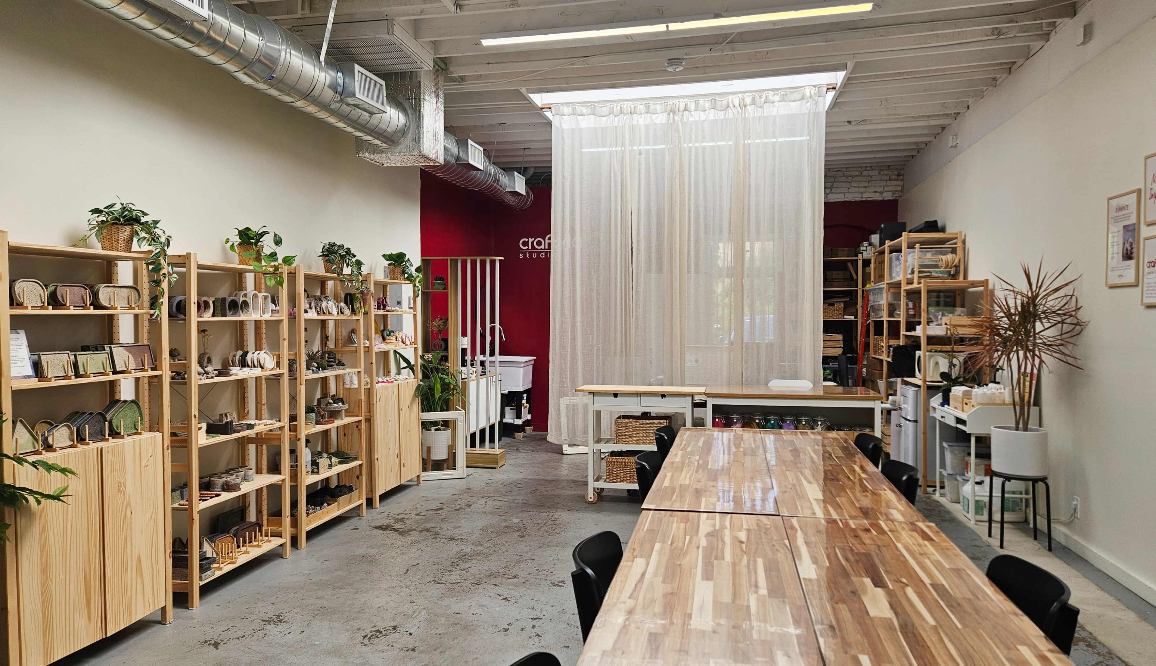 The image shows a spacious workshop area with wooden shelving filled with craft supplies, a large communal table, and a curtain dividing the space.