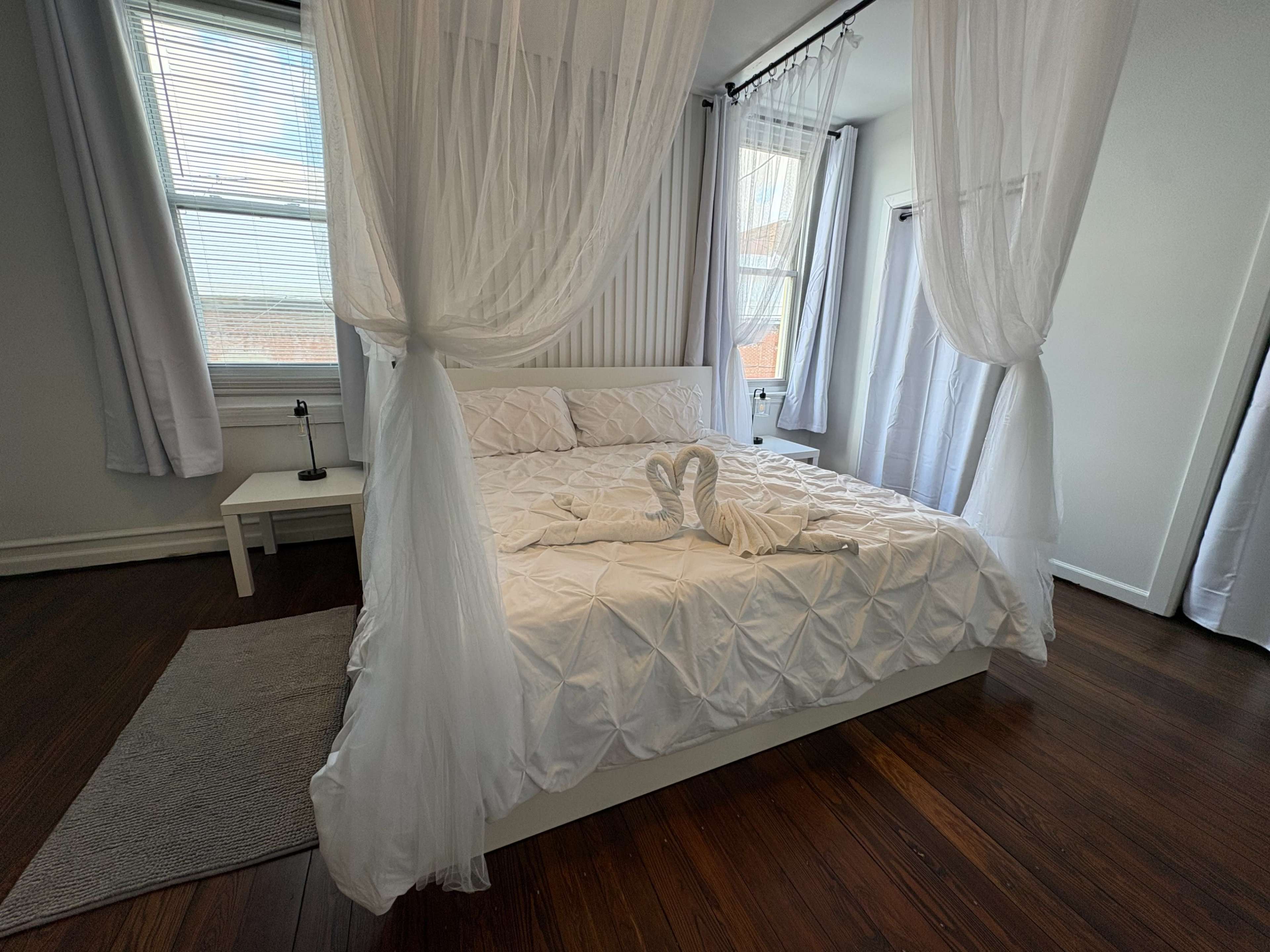 A neatly made bed with a white decorative cover is framed by sheer curtains in a bright, airy room with wooden floors.