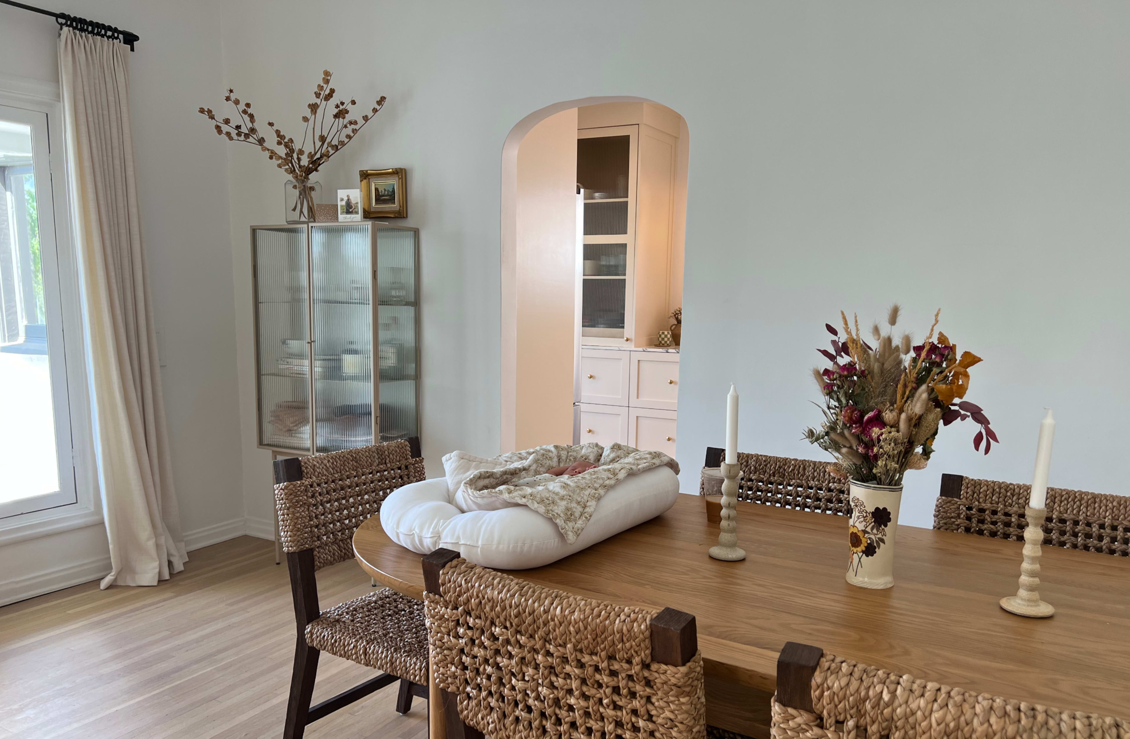A dining area features a wooden table surrounded by wicker chairs, with a decorative vase and a covered bowl at the center.