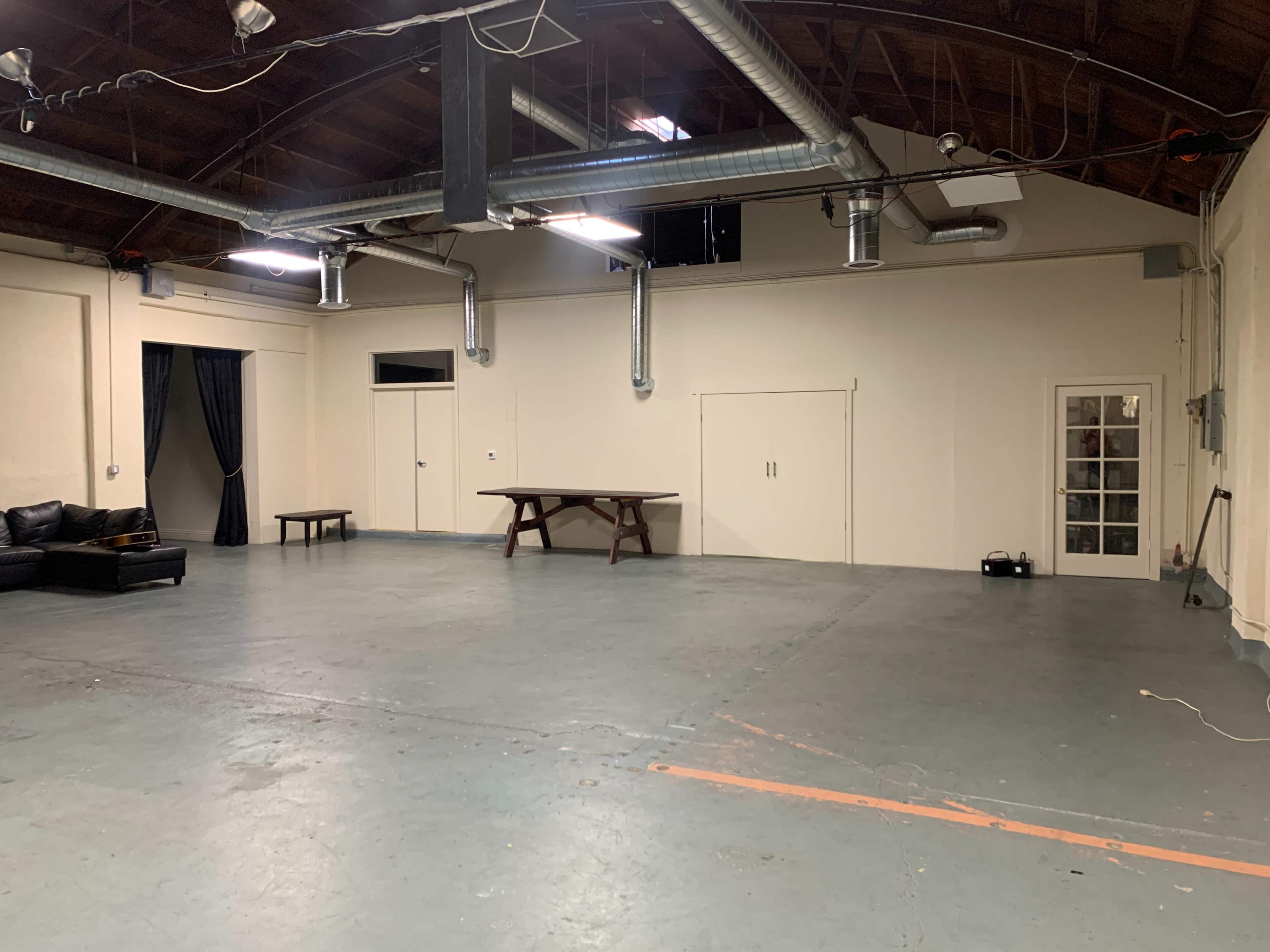 A spacious, unfurnished room with a table and a couch along one side, featuring industrial-style ventilation ducts on the ceiling.