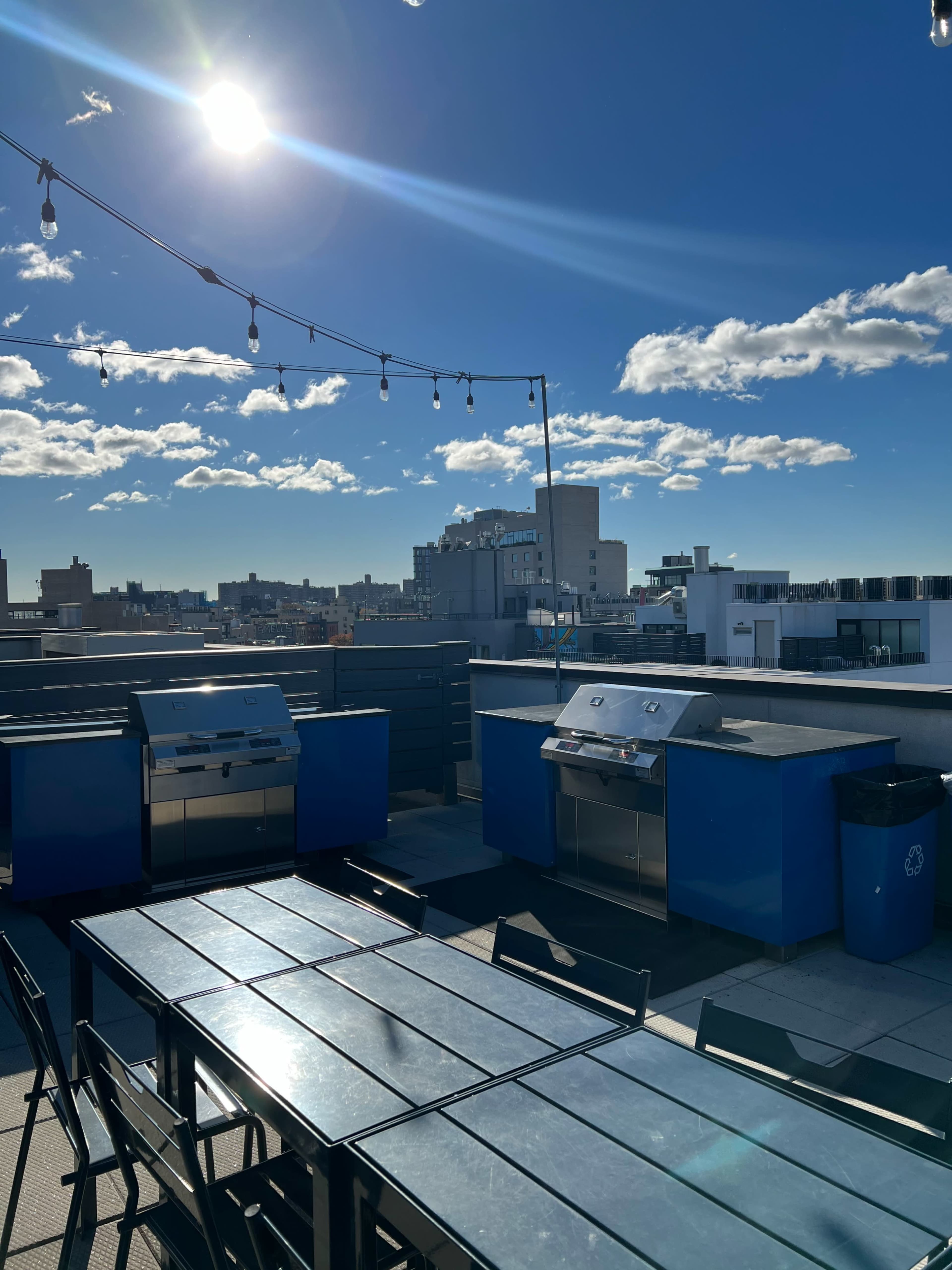 A rooftop grilling area features two blue grills and a long table under a bright sun and clear sky with scattered clouds.