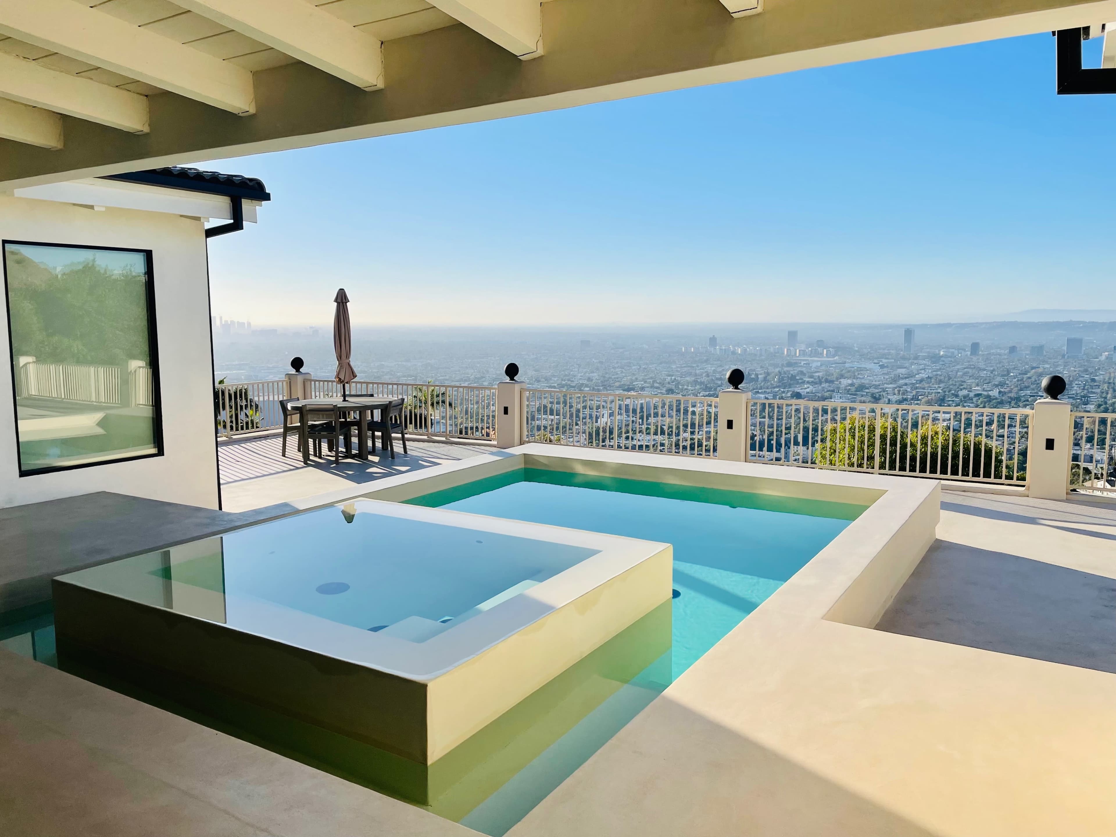 A modern patio features a pool and jacuzzi overlooking a city skyline with clear skies in the background.