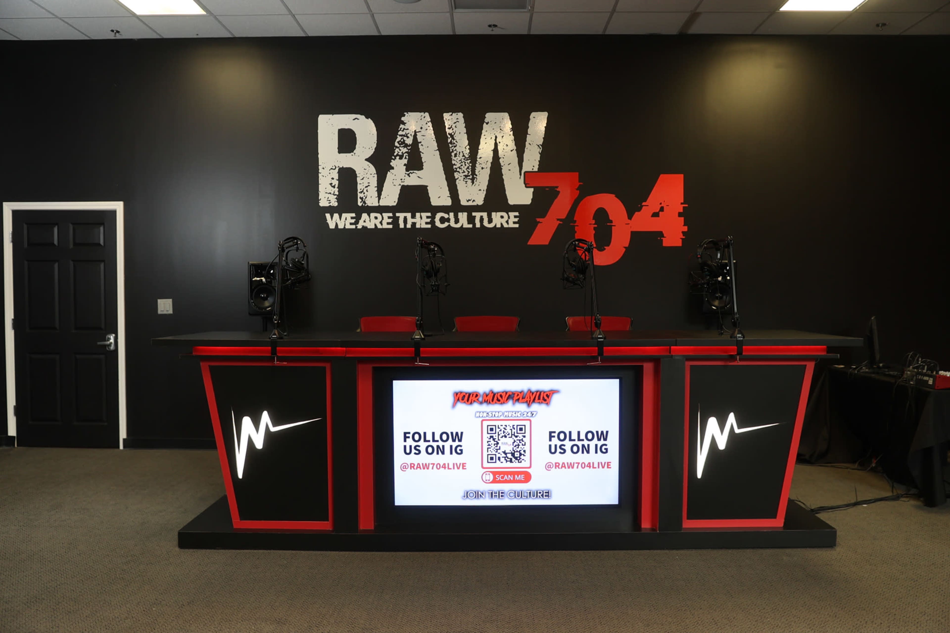 A modern broadcast studio setup with a large red and black counter, microphones, and a digital screen displaying social media handles against a black wall with the text "RAW 704."