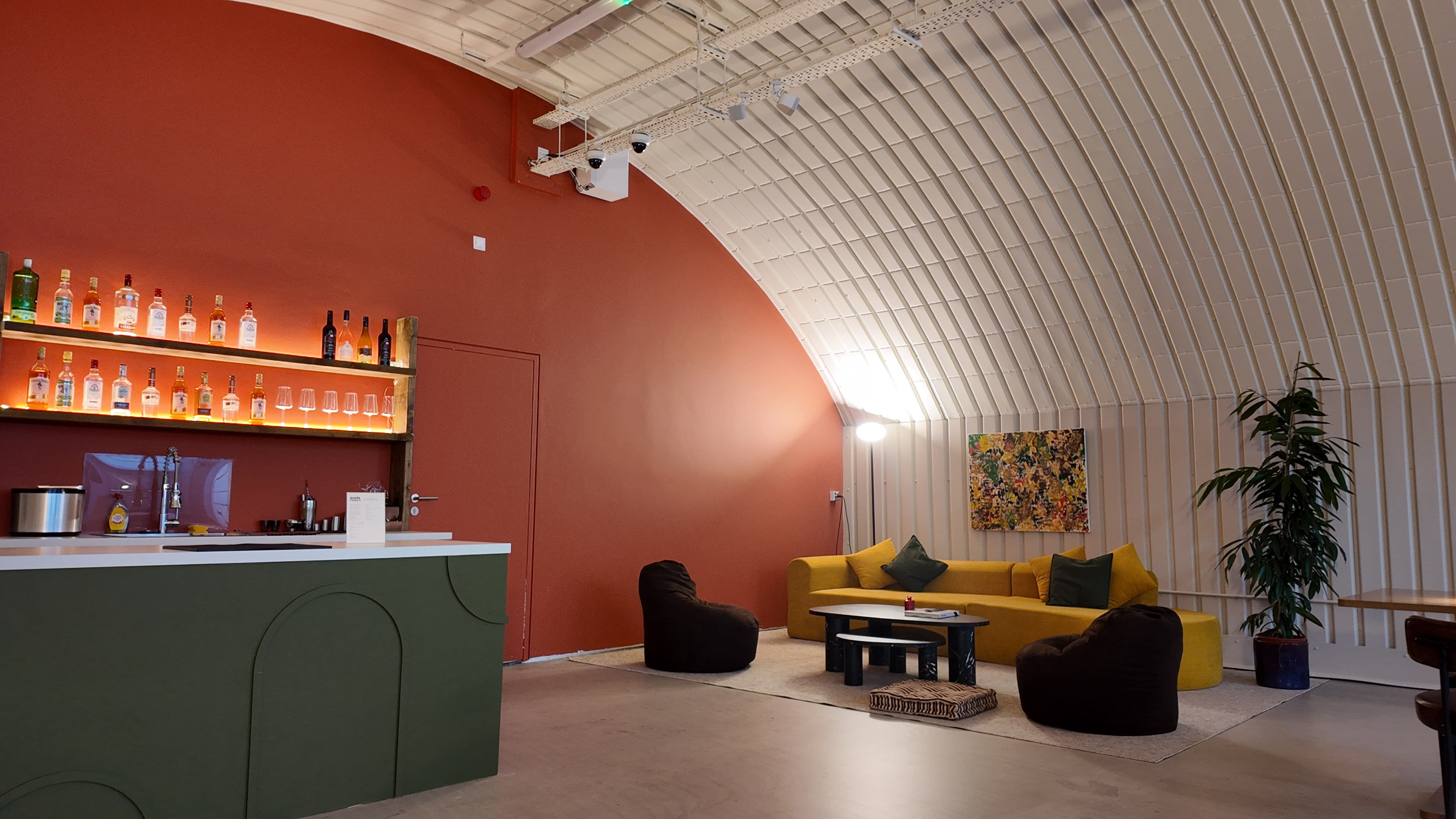 The image shows a modern lounge area with a bar, yellow couches, bean bag chairs, and a colorful artwork on the wall, all under a curved ceiling.