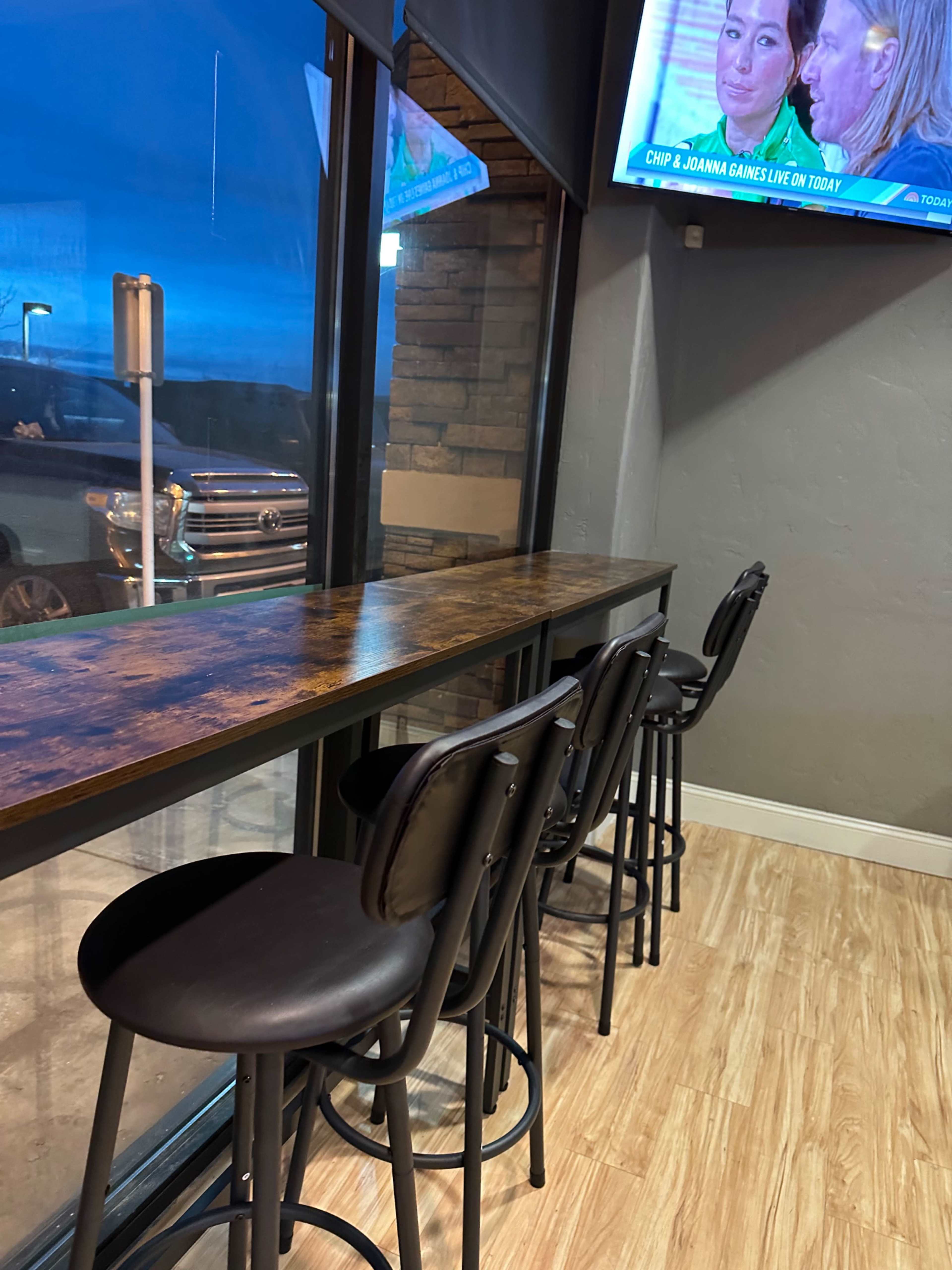The image shows a row of black barstools lined up against a wooden counter by a large window, with a television displaying a show in the background.