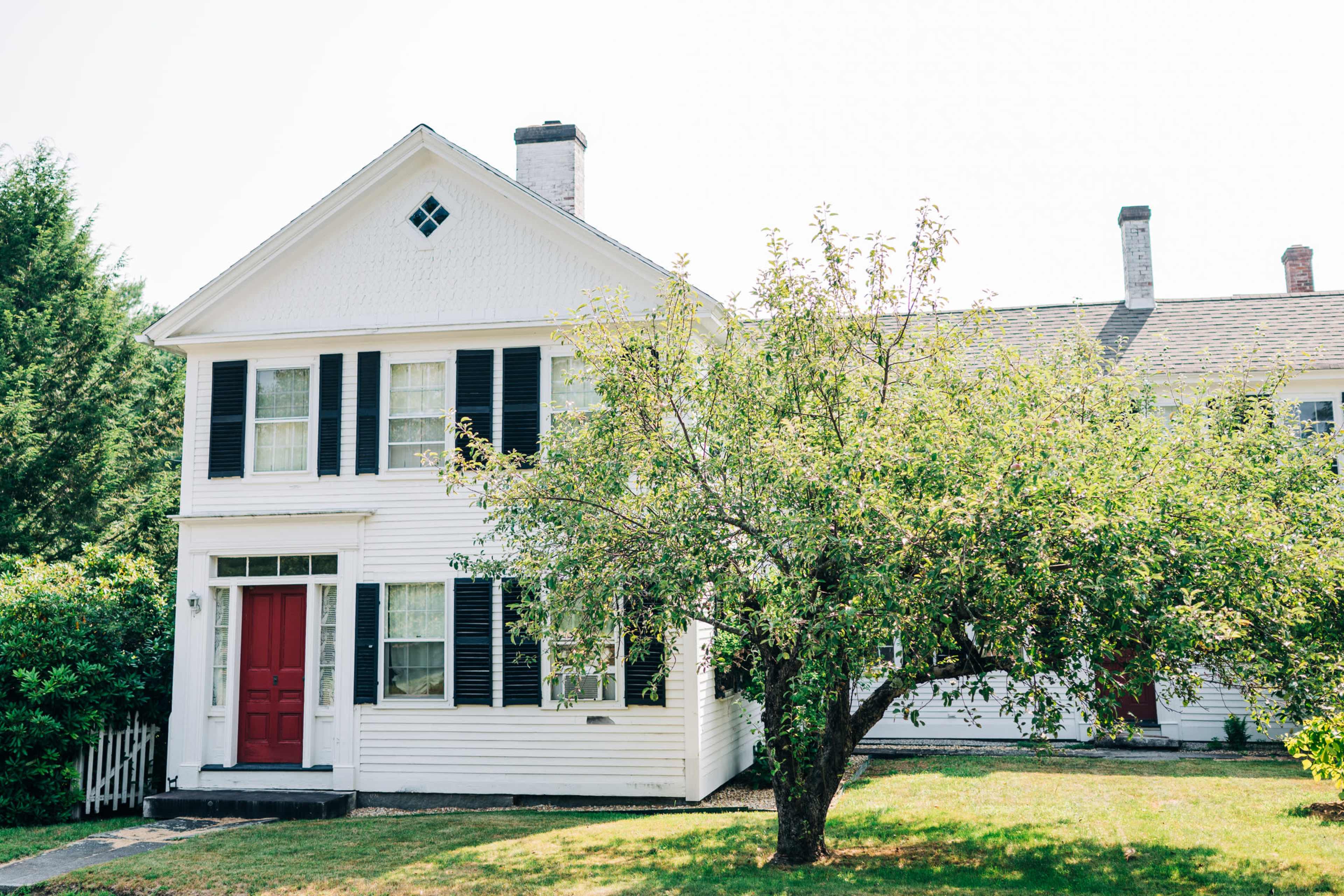 A white two-story house with black shutters and a red front door is situated next to a grassy yard with a tree in the foreground.