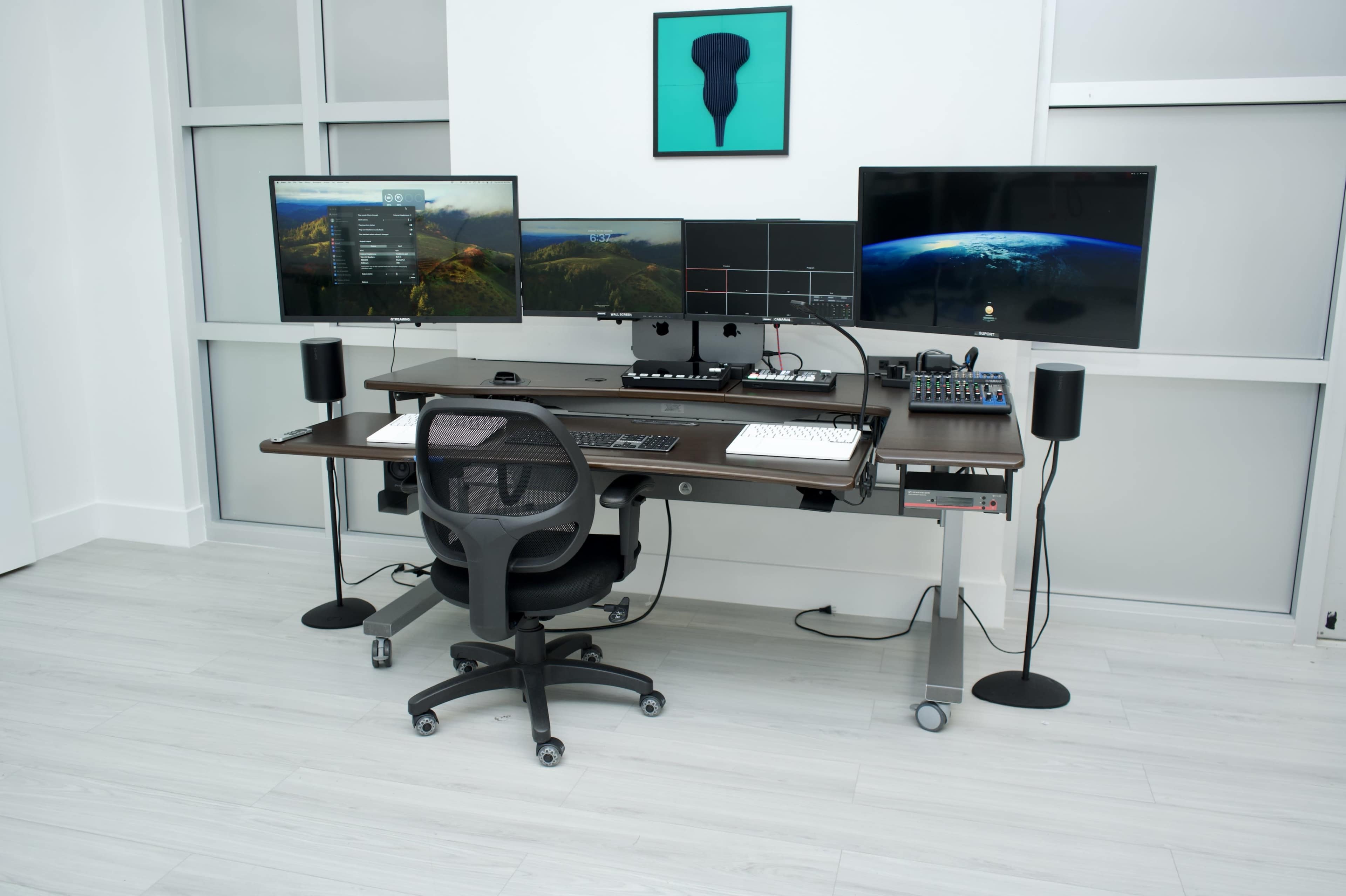 The image shows a modern workspace featuring a large desk with dual monitors, a keyboard, a mouse, and audio equipment, accompanied by an ergonomic chair and floor speakers.