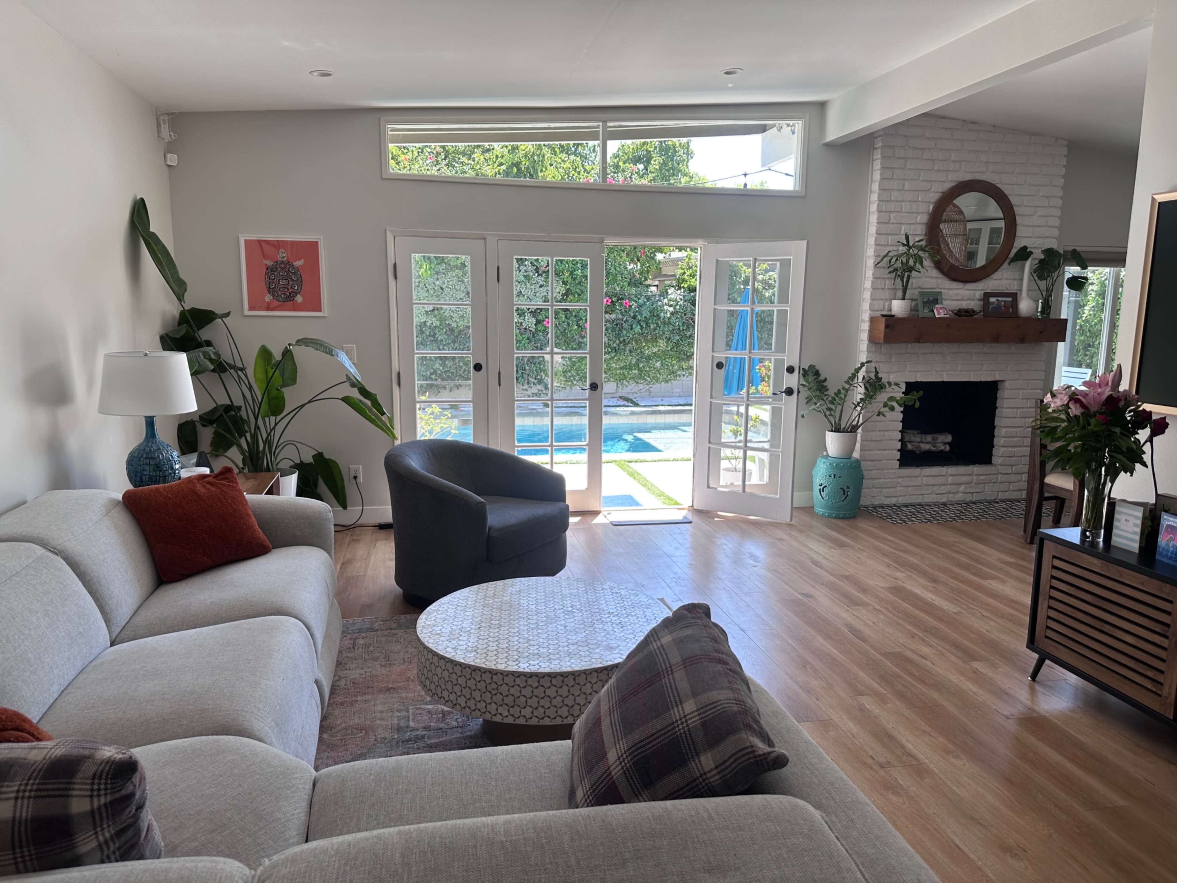 The image shows a modern living room with a light gray sectional sofa, a round coffee table, a decorative fireplace, and large glass doors leading to an outdoor area.