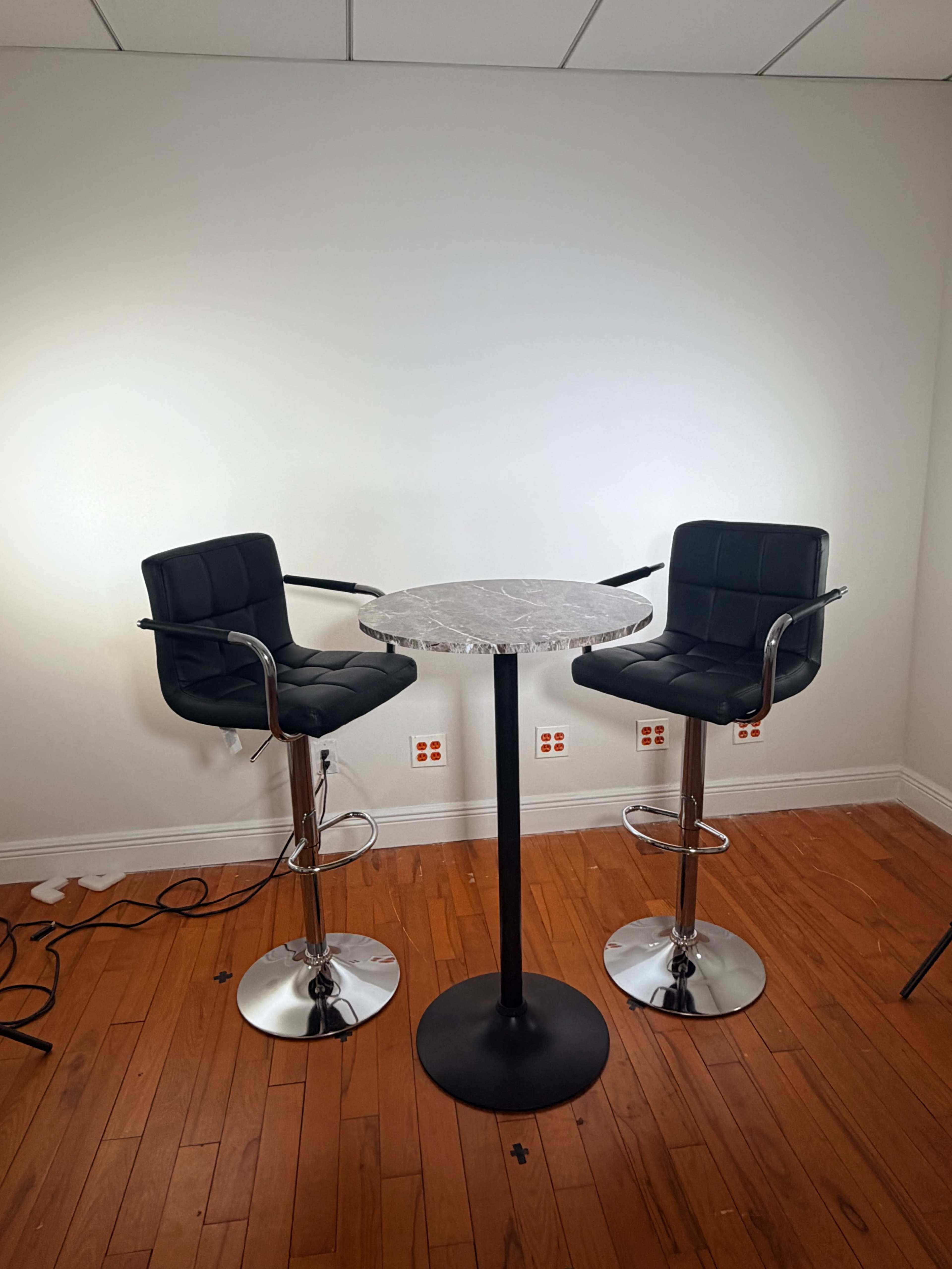 Two black bar stools are positioned around a circular marble-top table in a room with wood flooring and white walls.