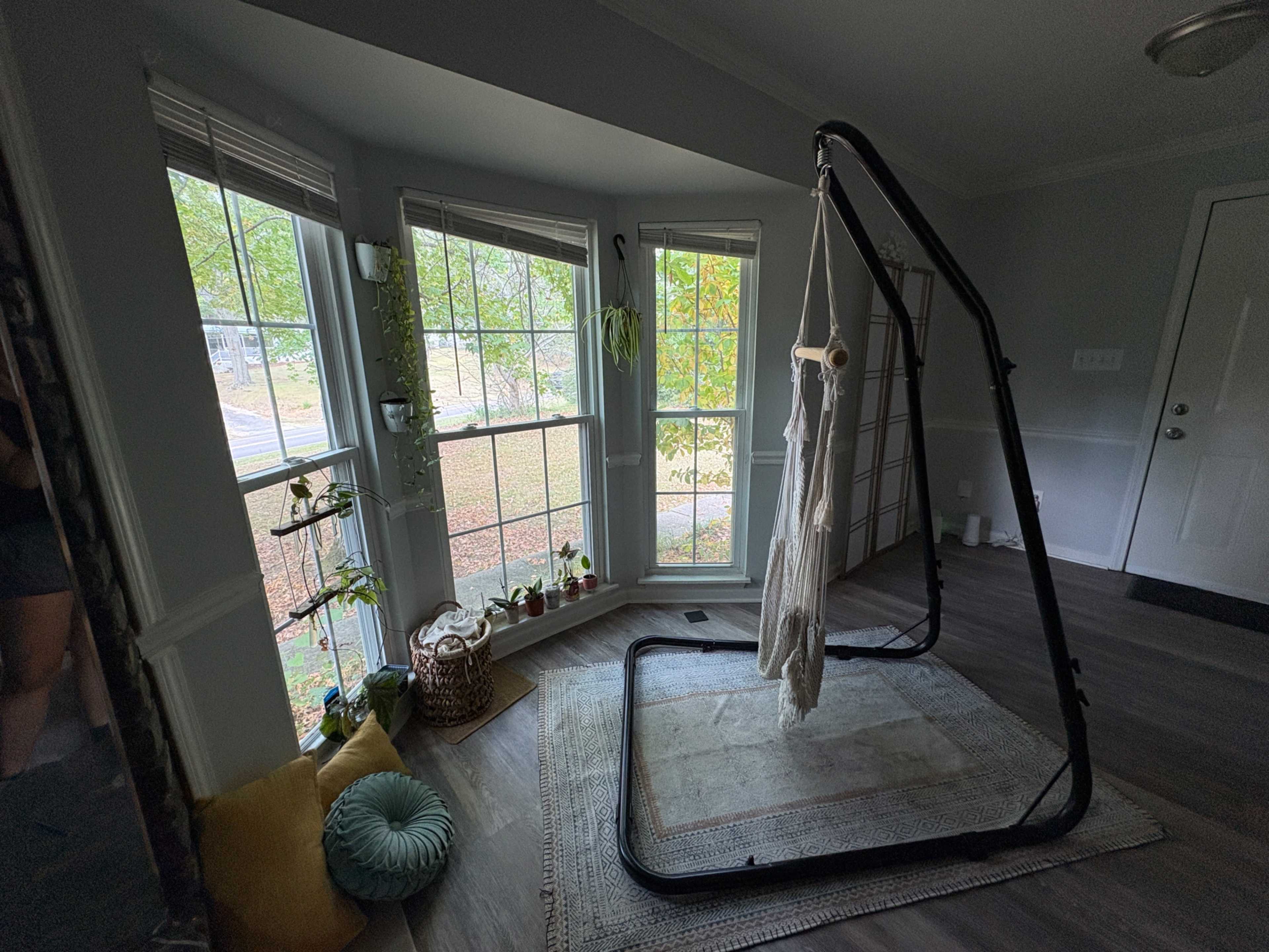 A macrame swing hangs in a well-lit corner of a room with large windows overlooking a tree-filled view.
